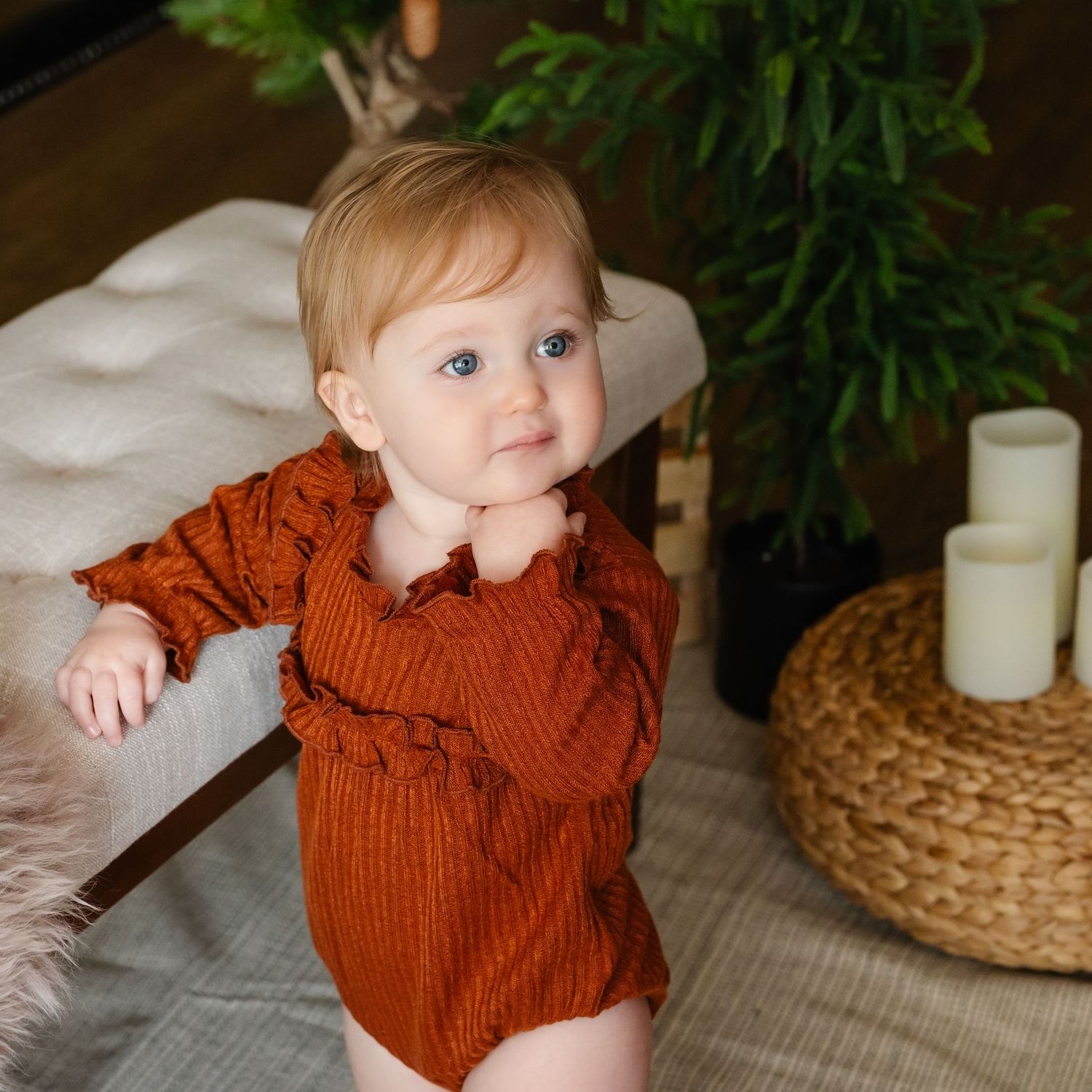 A baby in a rust-colored outfit leans on a table.