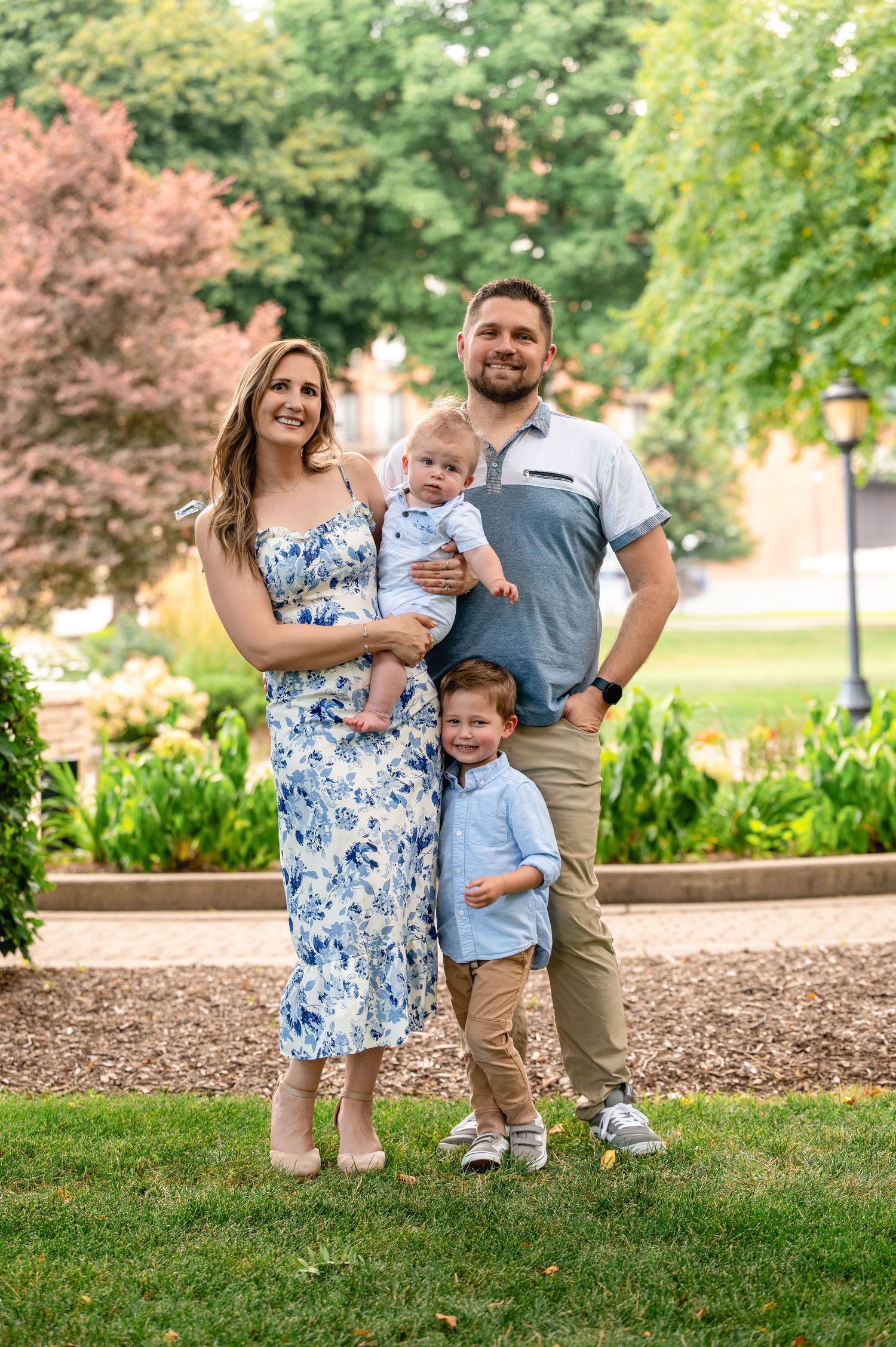Family of four posing together in park with lush greenery and flowering trees.