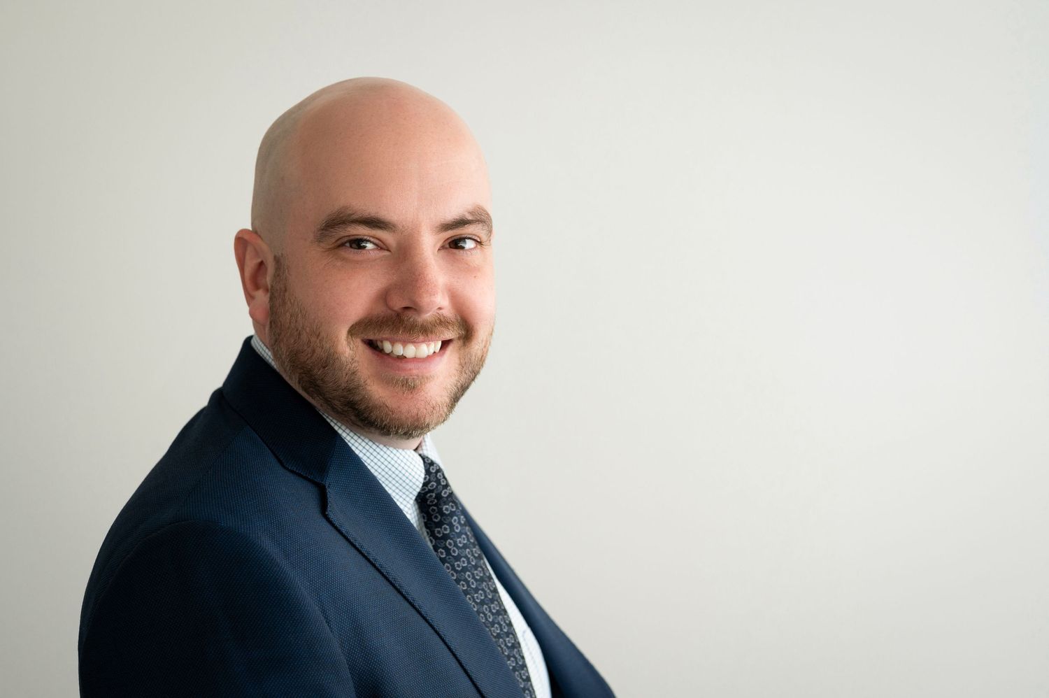 Professional headshot of person in navy suit against light gray background.
