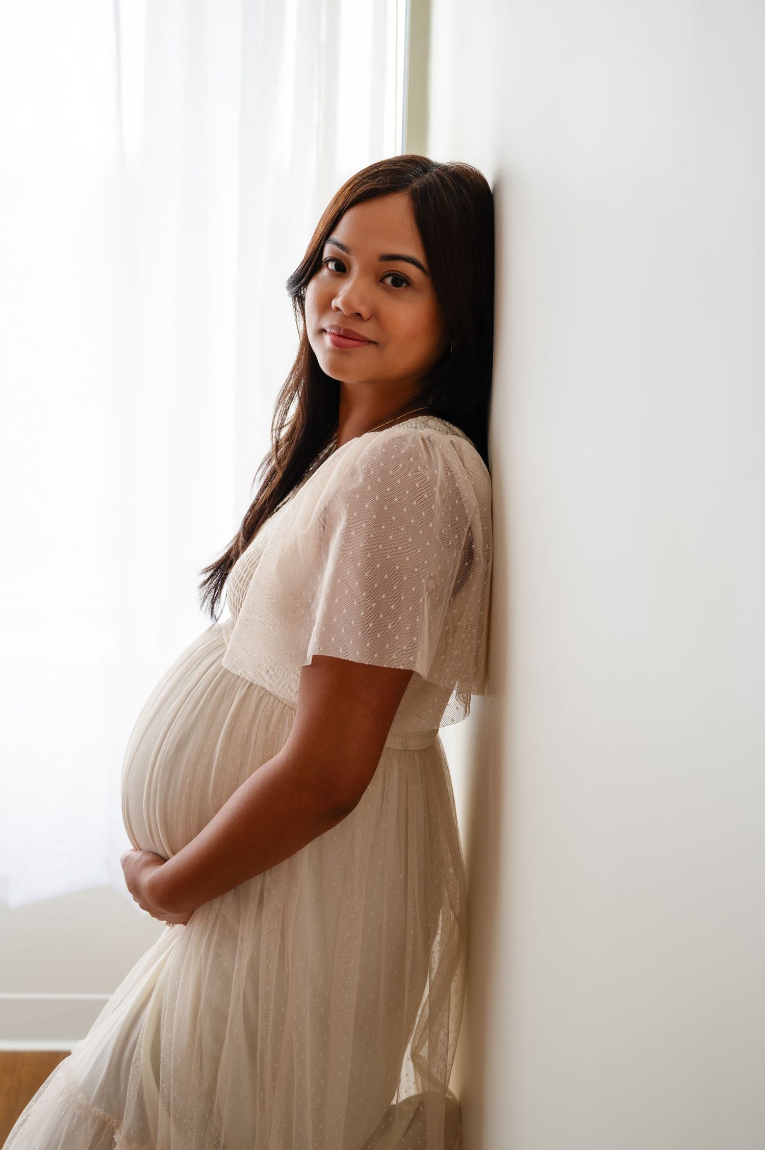 Maternity portraits showing expectant mother in white dress against bright window.