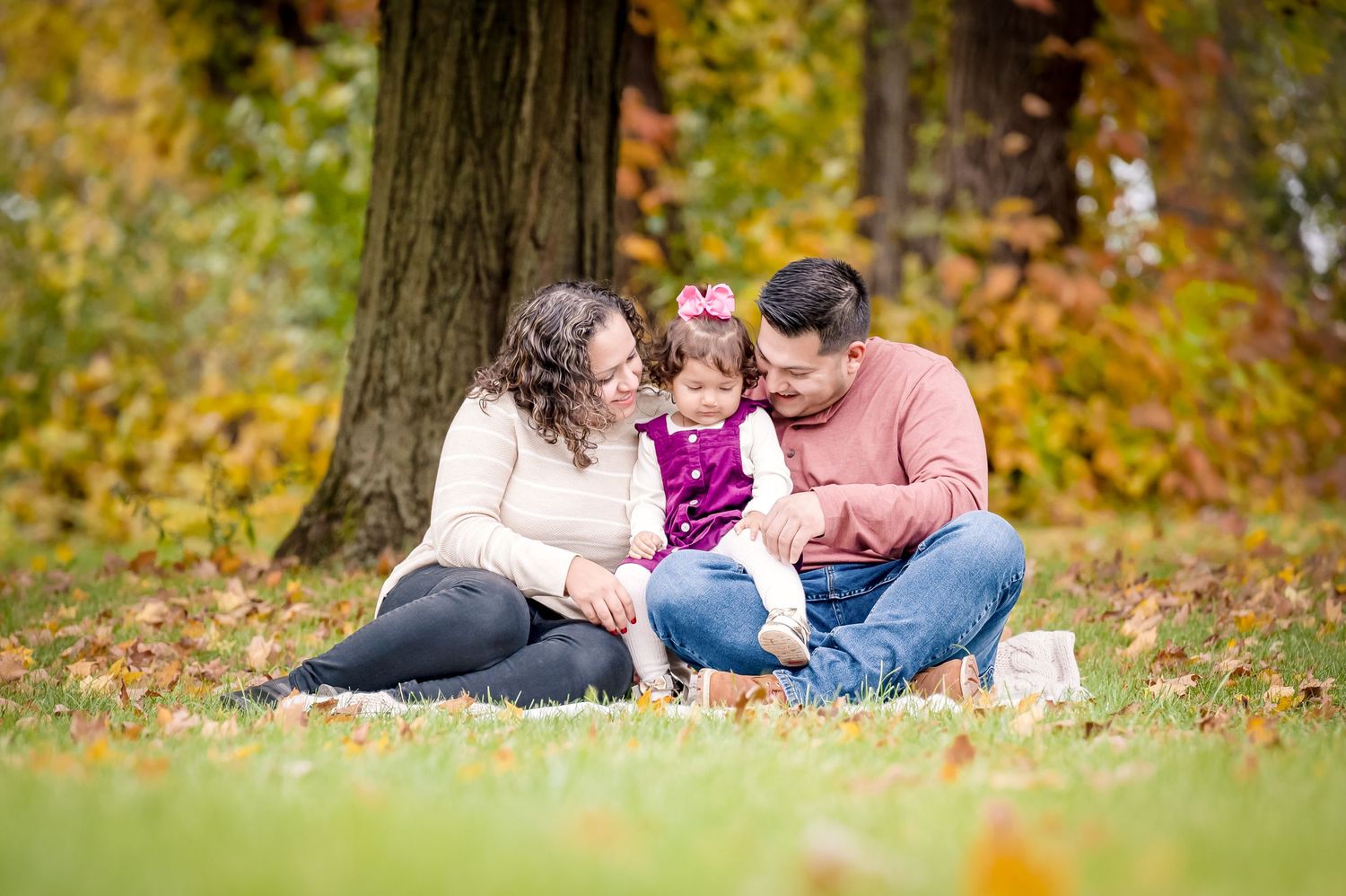 A family with a young toddler snuggle in this tender moment with beautiful fall foliage in pops of orange, yellow and red surround them.