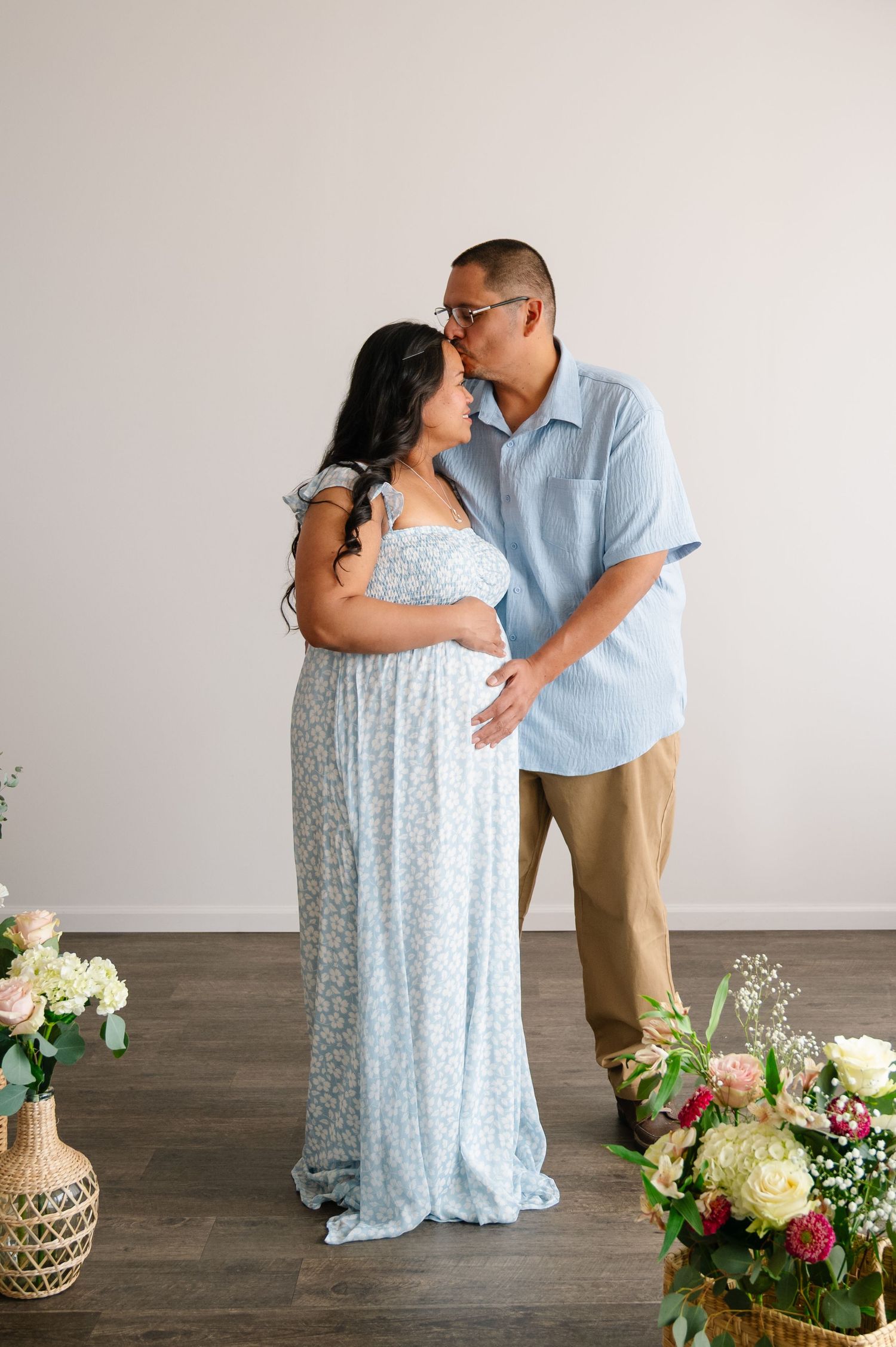 A romantic maternity portrait of a couple in light colored clothing against a white backdrop.