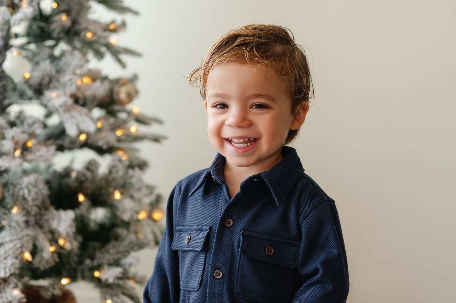 Child in navy blue outfit smiles brightly in front of Christmas tree.