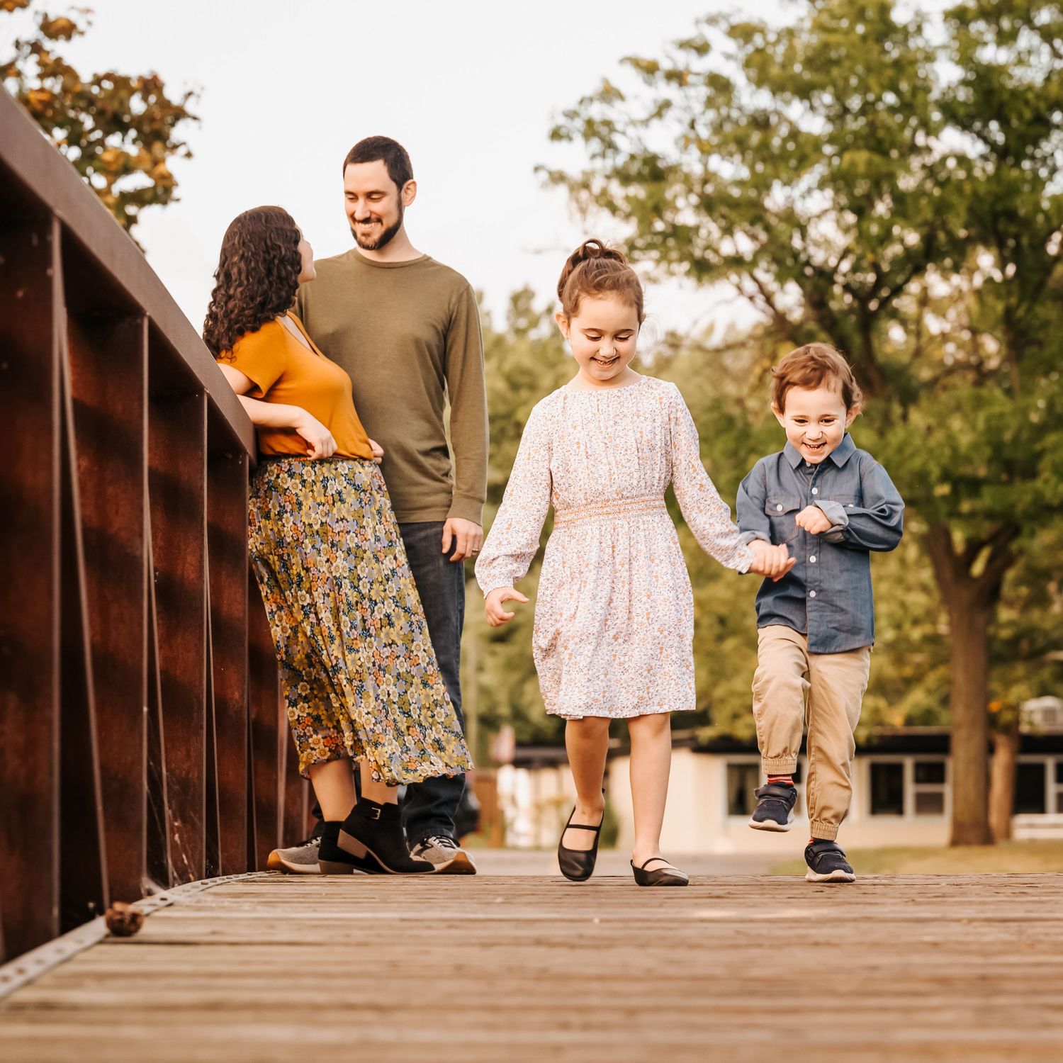 A family walks together holding hands across a wooden bridge in an autumn setting.