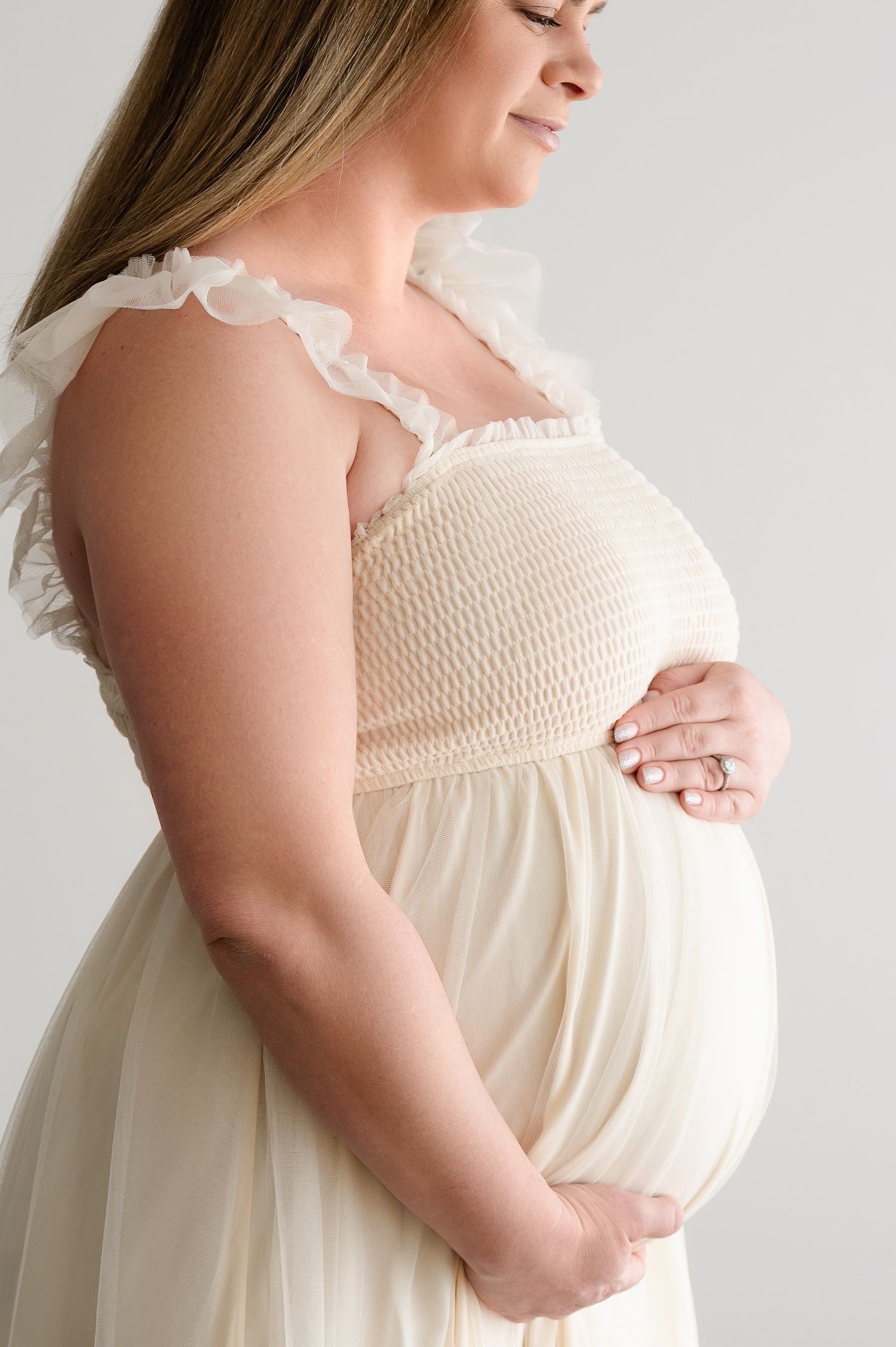A side view of an expectant mother wearing a cream-colored smocked dress with ruffled details.
