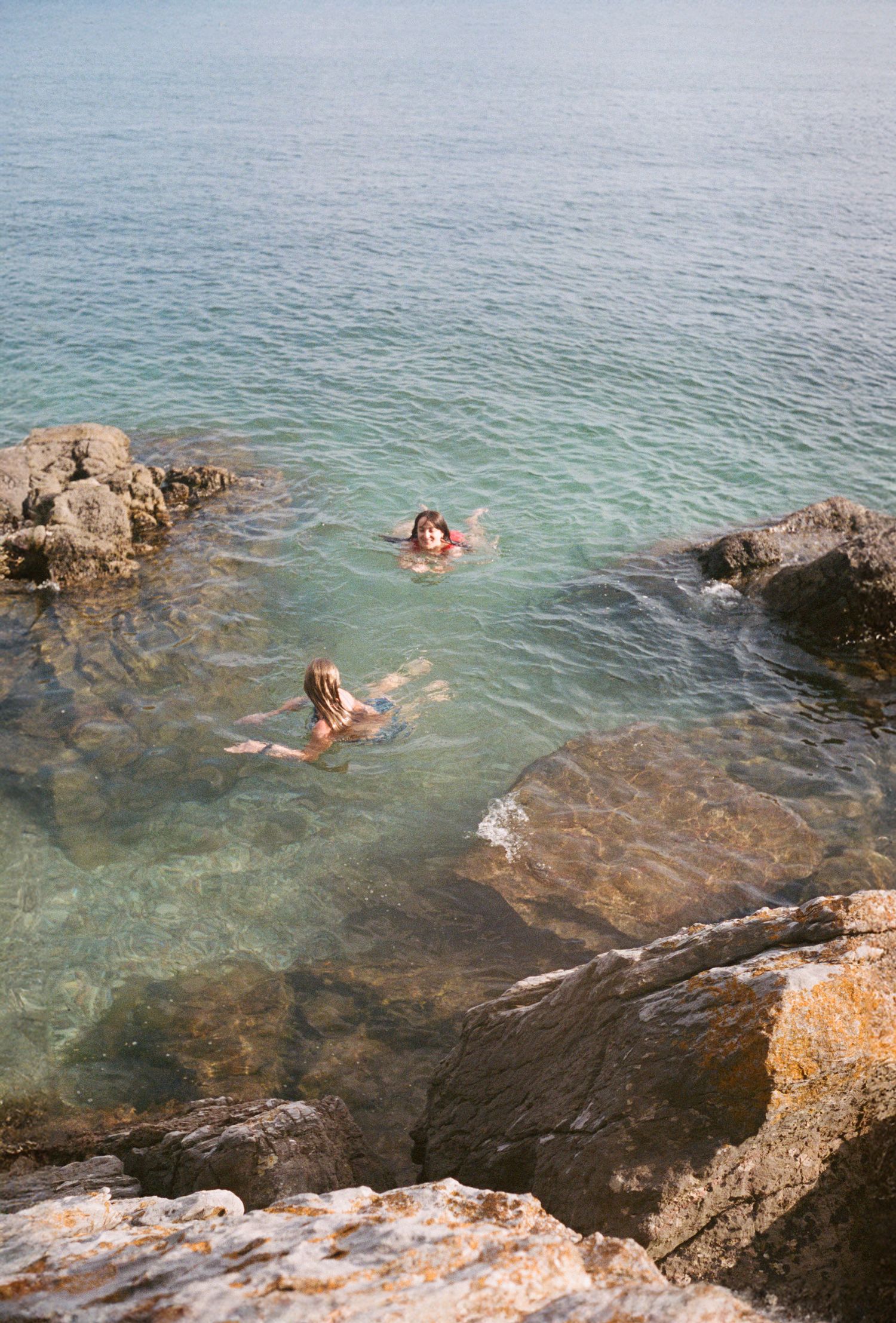 Two people swimming in crystal clear turquoise waters between rocky coastal cliffs.