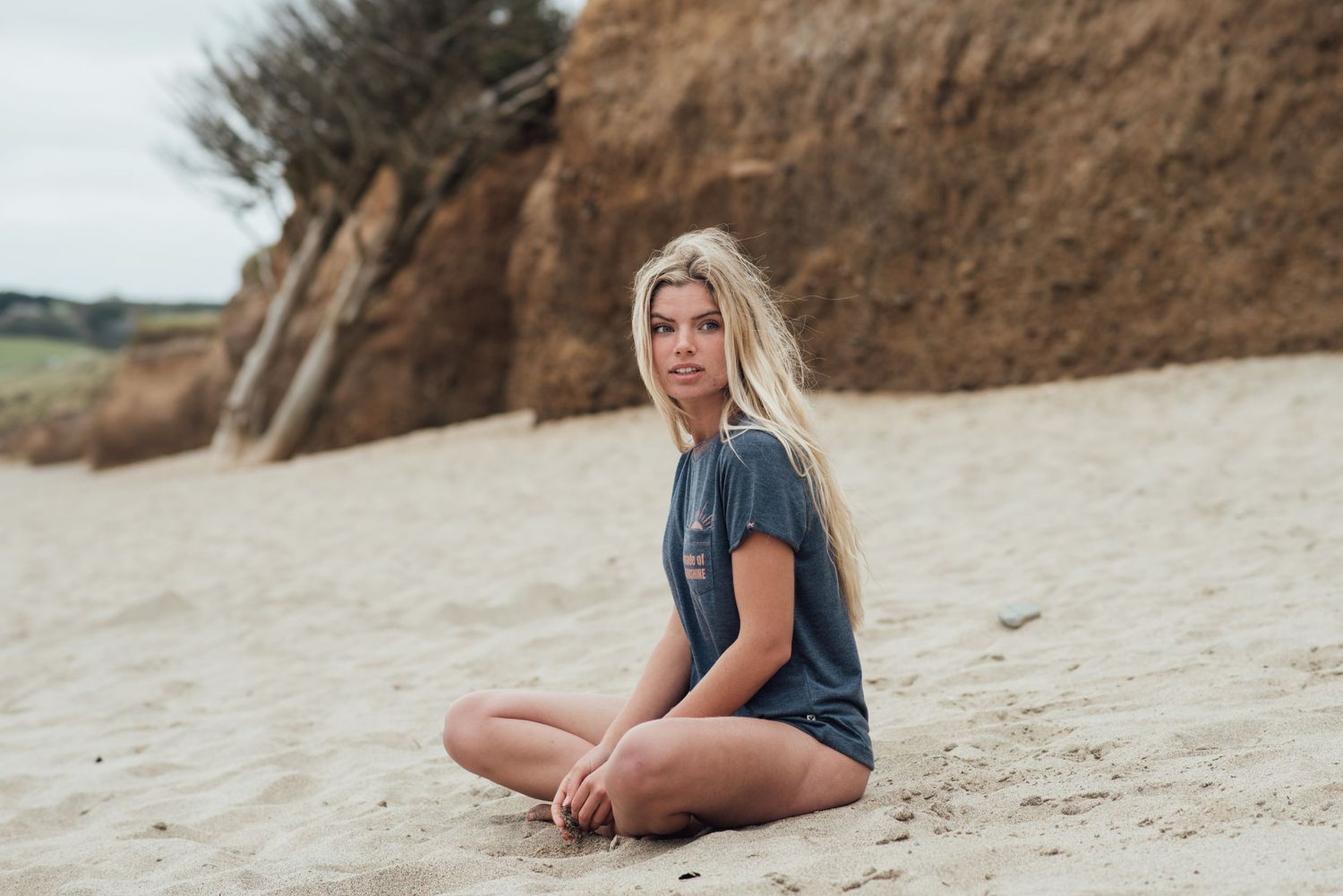 Someone sitting casually on a sandy beach wearing a dark t-shirt near a coastal cliff.