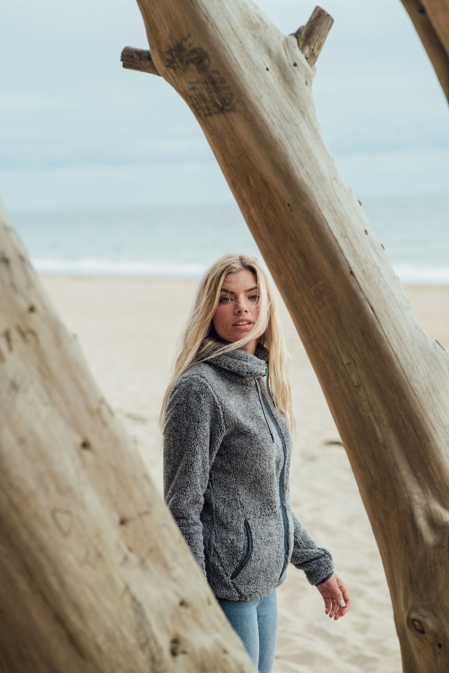 Person in grey sweater standing against driftwood on a cloudy beach.