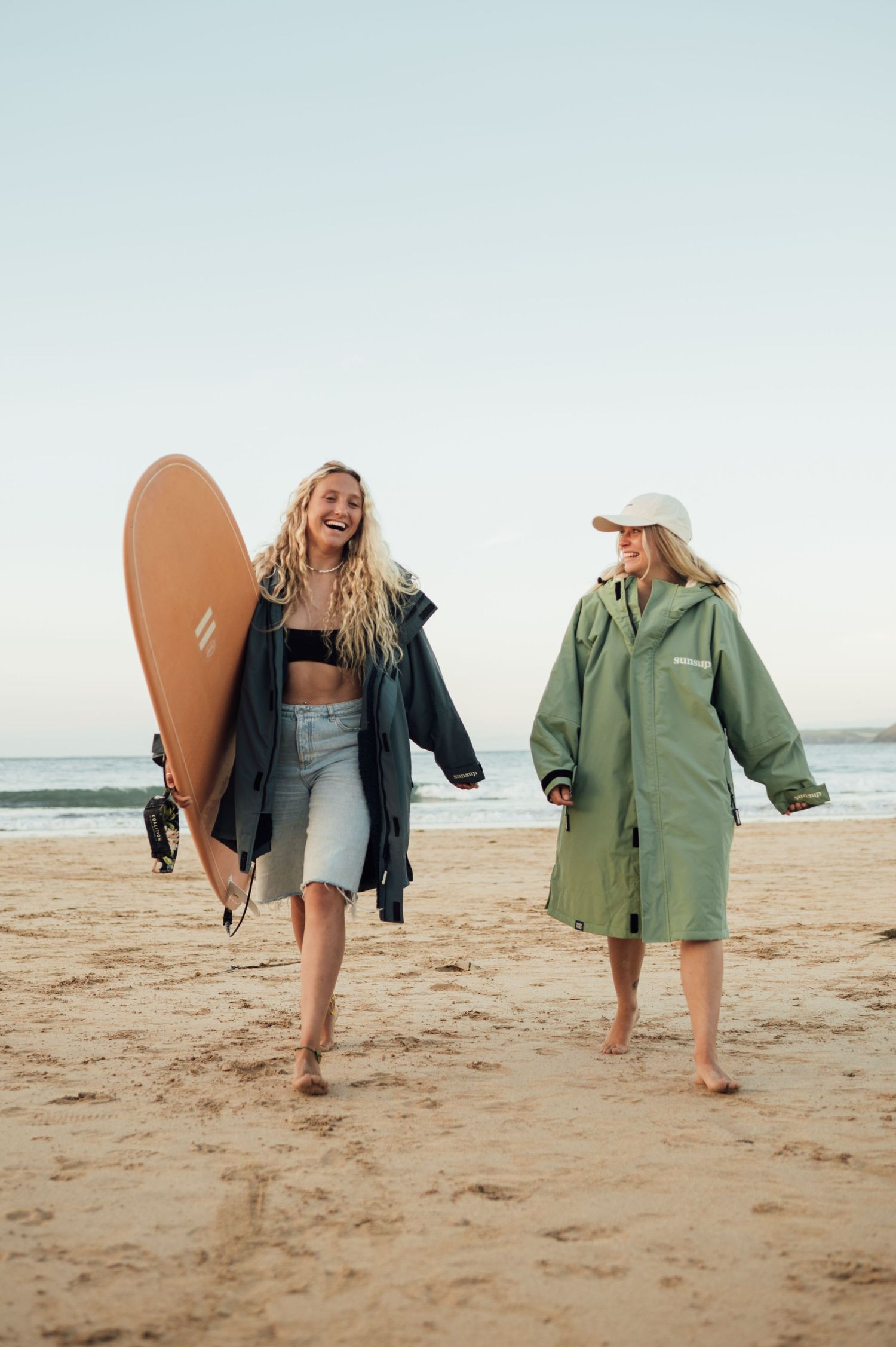 Two surfers stroll along the beach with surfboards and jackets during sunset.
