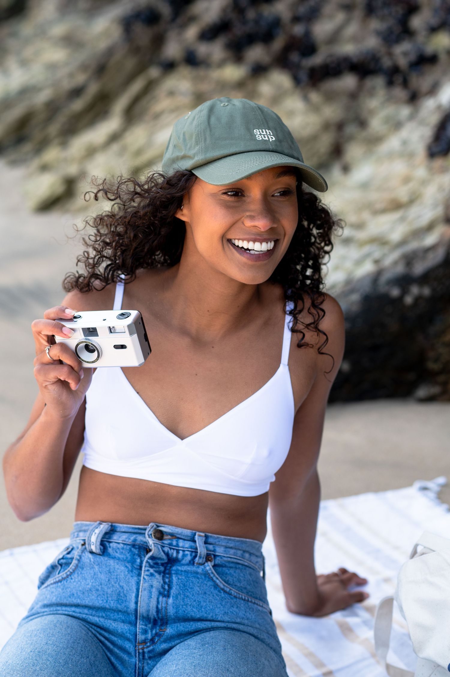 Someone wearing a green cap and white crop top holds a vintage camera while sitting on a beach.