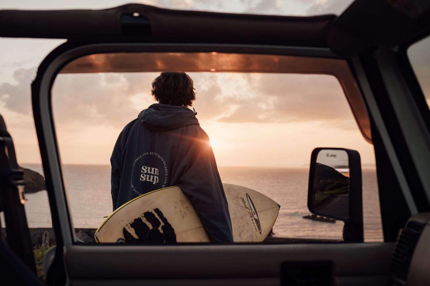 Silhouette of a surfer watching sunset through a car window while holding a surfboard.