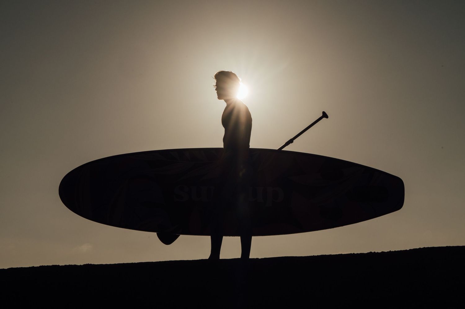 Silhouette of surfer holding board against bright sun on beach hill.