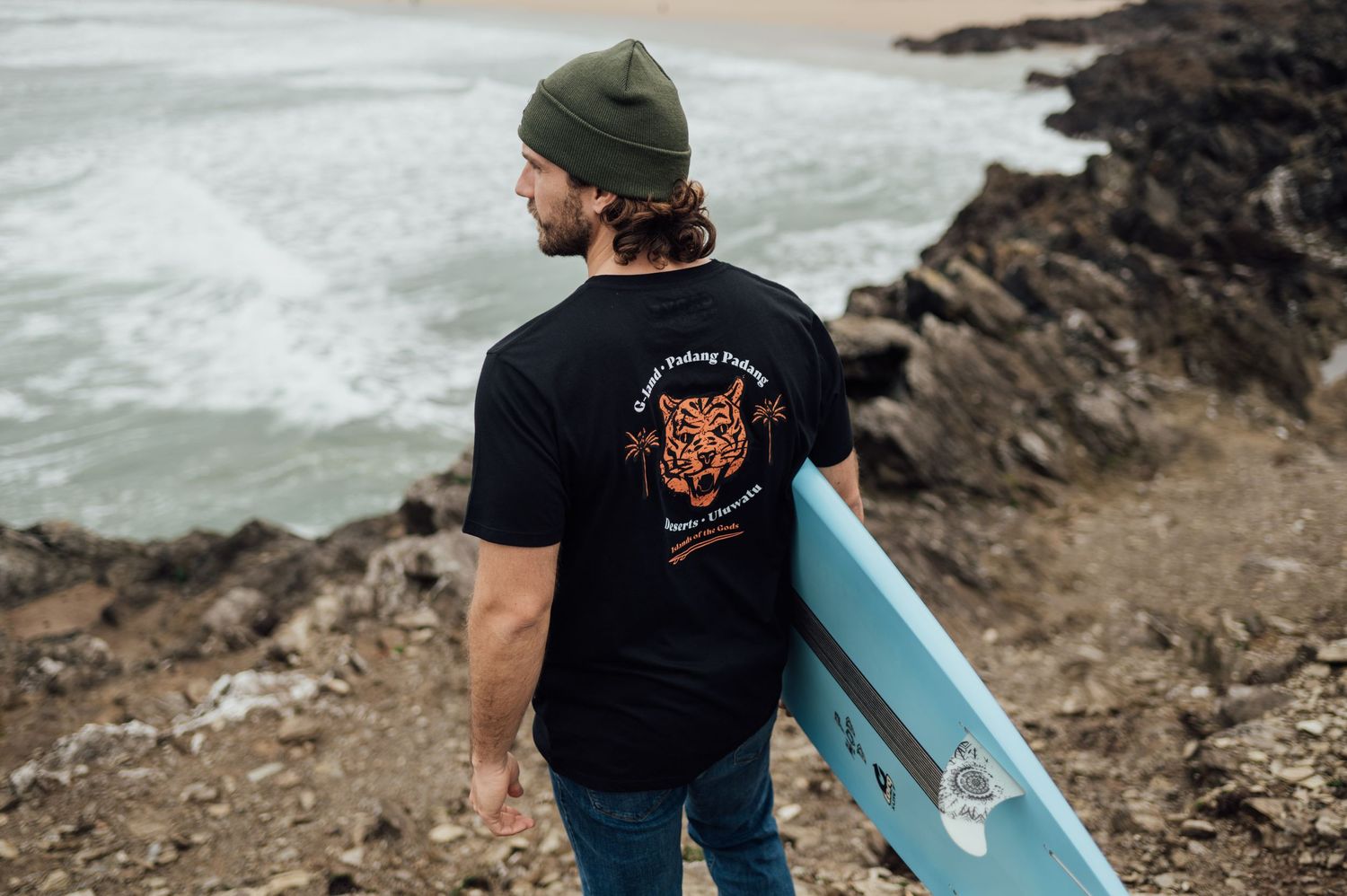 Surfer with tiger graphic t-shirt holding blue surfboard on rocky coastal cliff.