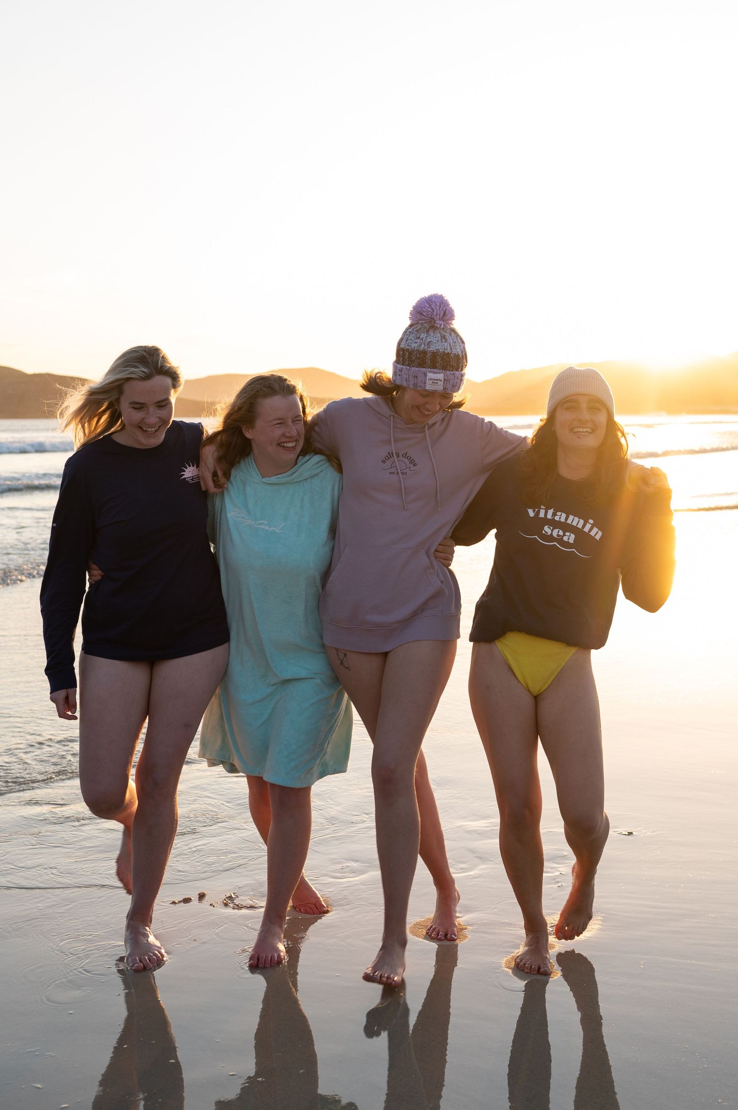 A group of friends in casual beachwear walking together on the sand at sunset.