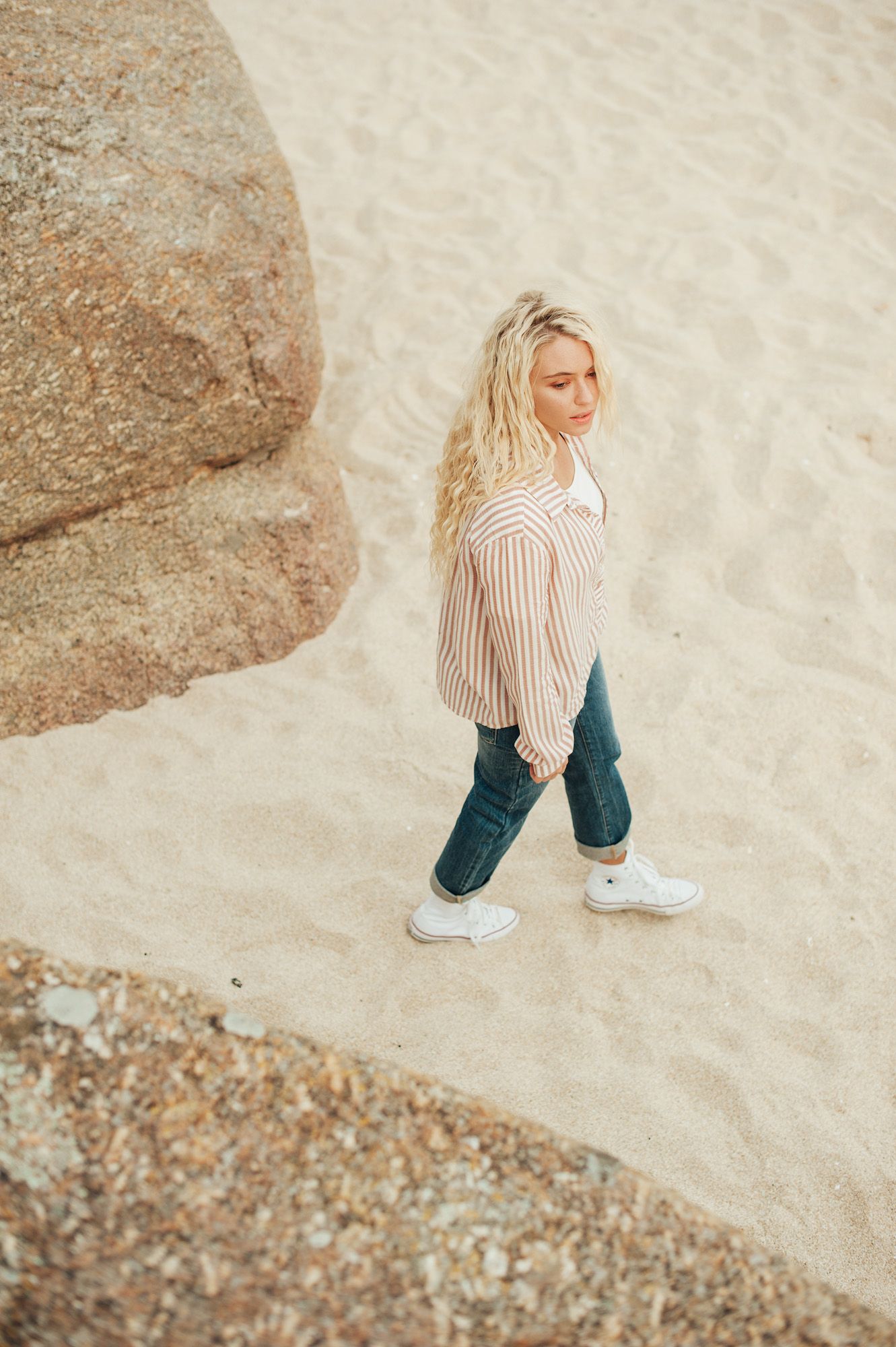 Someone walking on beach sand wearing a striped shirt and jeans between large coastal rocks.