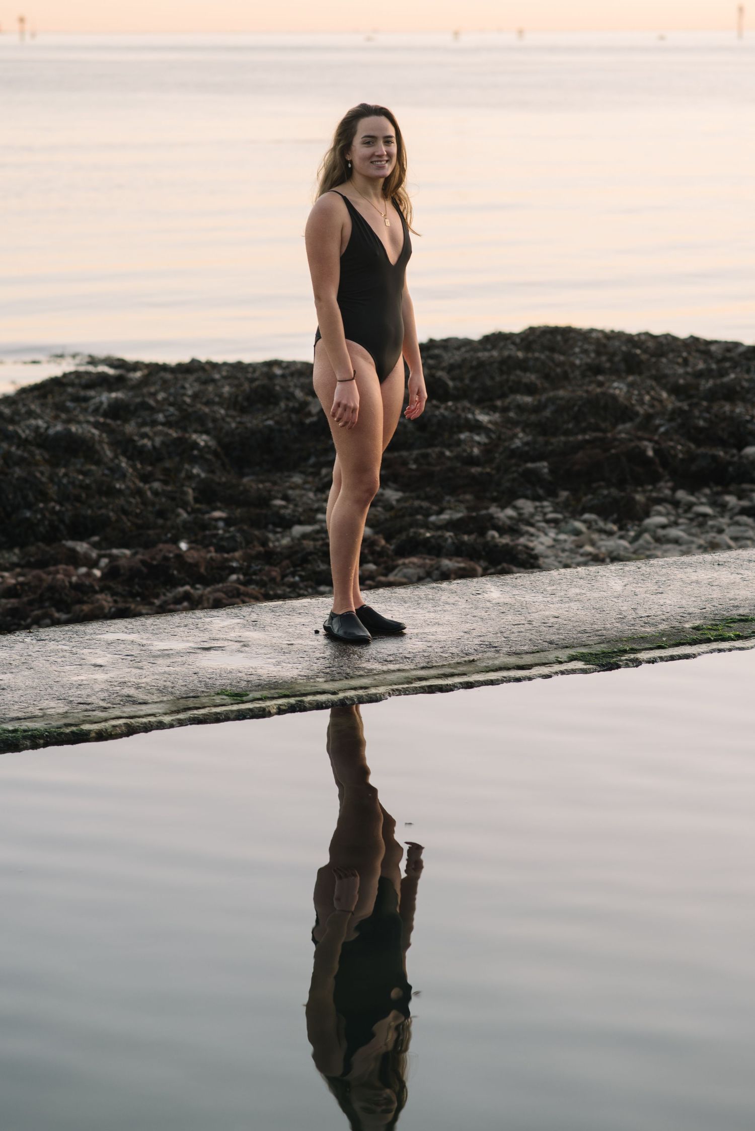 Someone in a black swimsuit standing on rocks by the ocean with their reflection in still water.