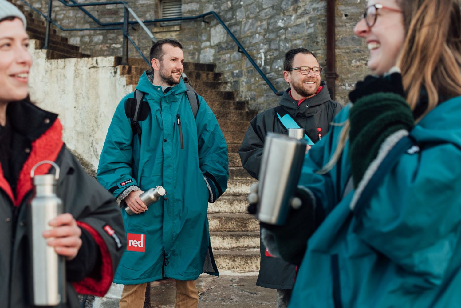 Group of friends in teal coats sharing drinks and conversation outdoors. Dryrobe. Changing robes