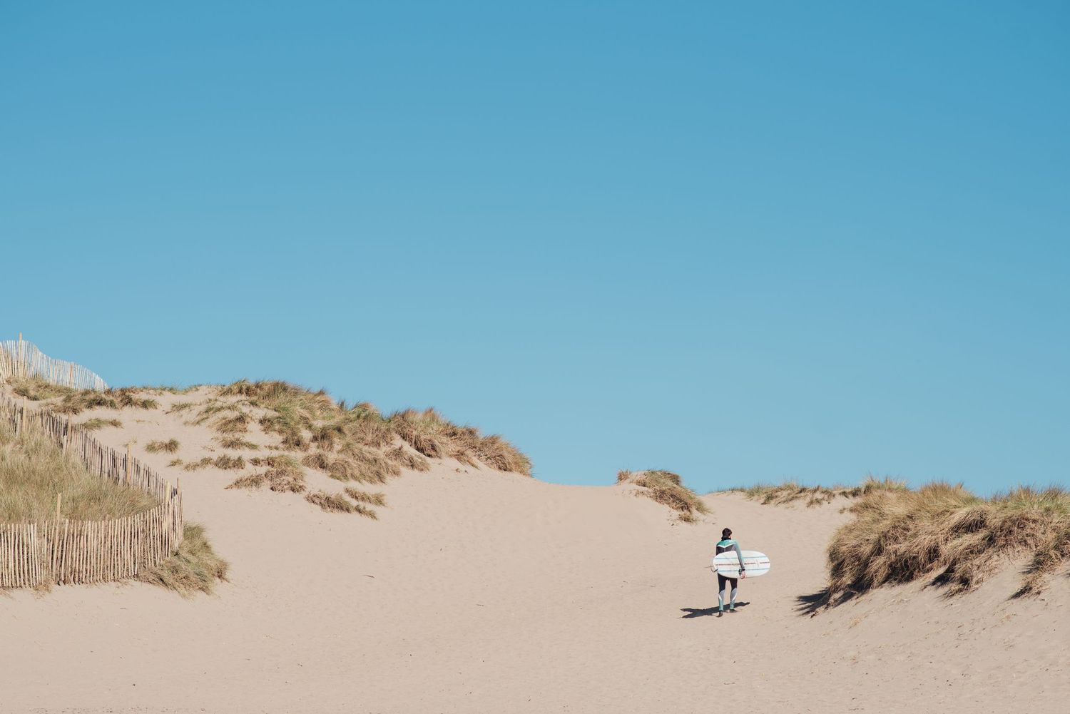 Lone figure surfer walking across vast sand dunes under bright blue sky.
