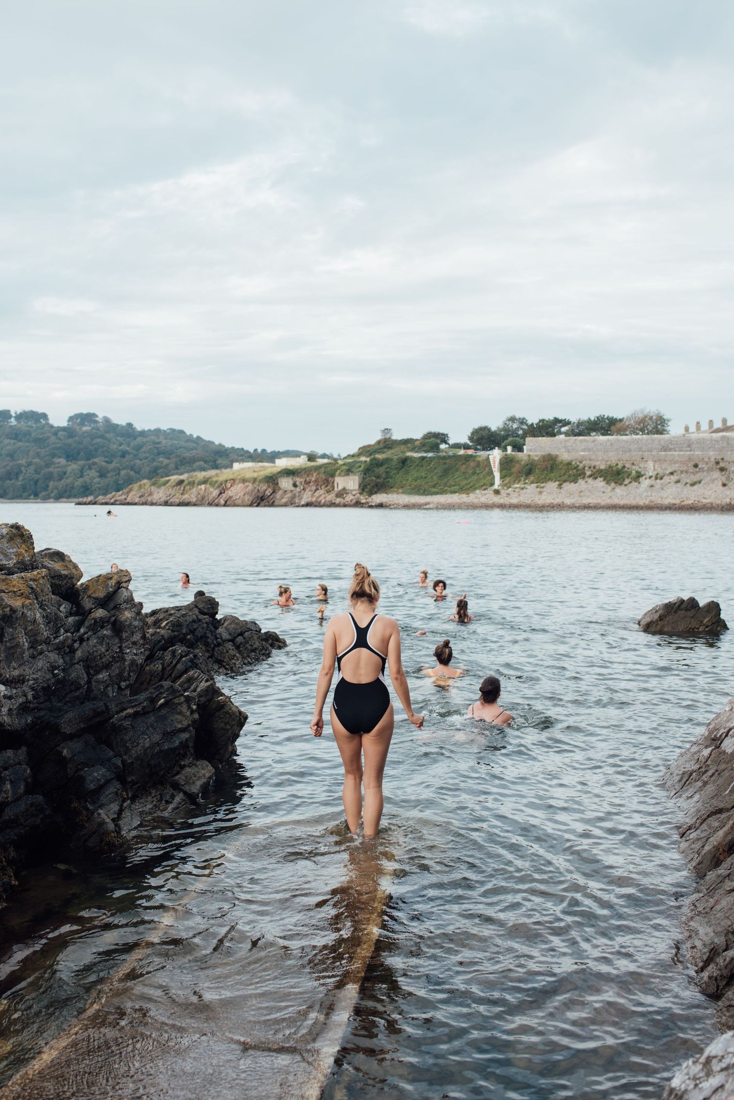 Scene of swimmers wading into rocky coastal waters on an overcast day.