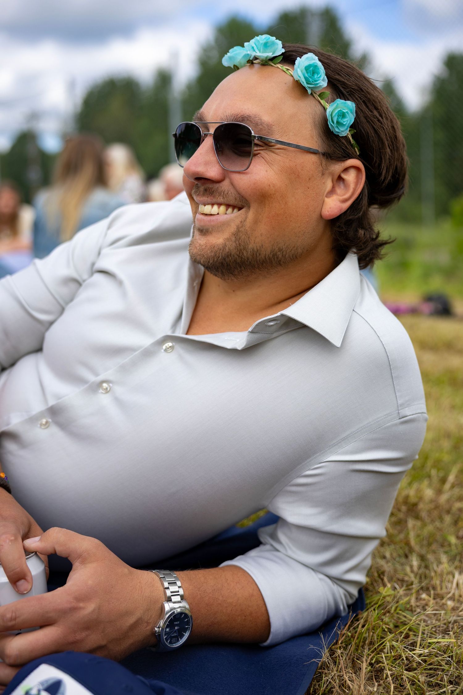 Person som kopplar av på gräset iklädd vit skjorta och blå blomsterkrans på en solig dag.