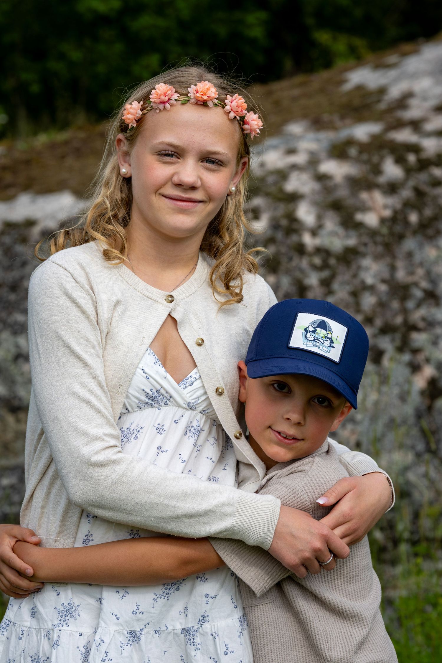 Barn omfamnar varandra utomhus mot en stenig bakgrund, klädda i vita kläder och blommiga accessoarer.