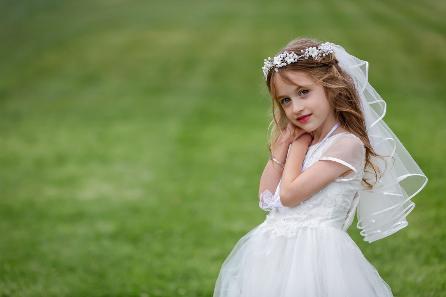 A young child in a white dress and floral crown with veil poses on green grass outdoors.