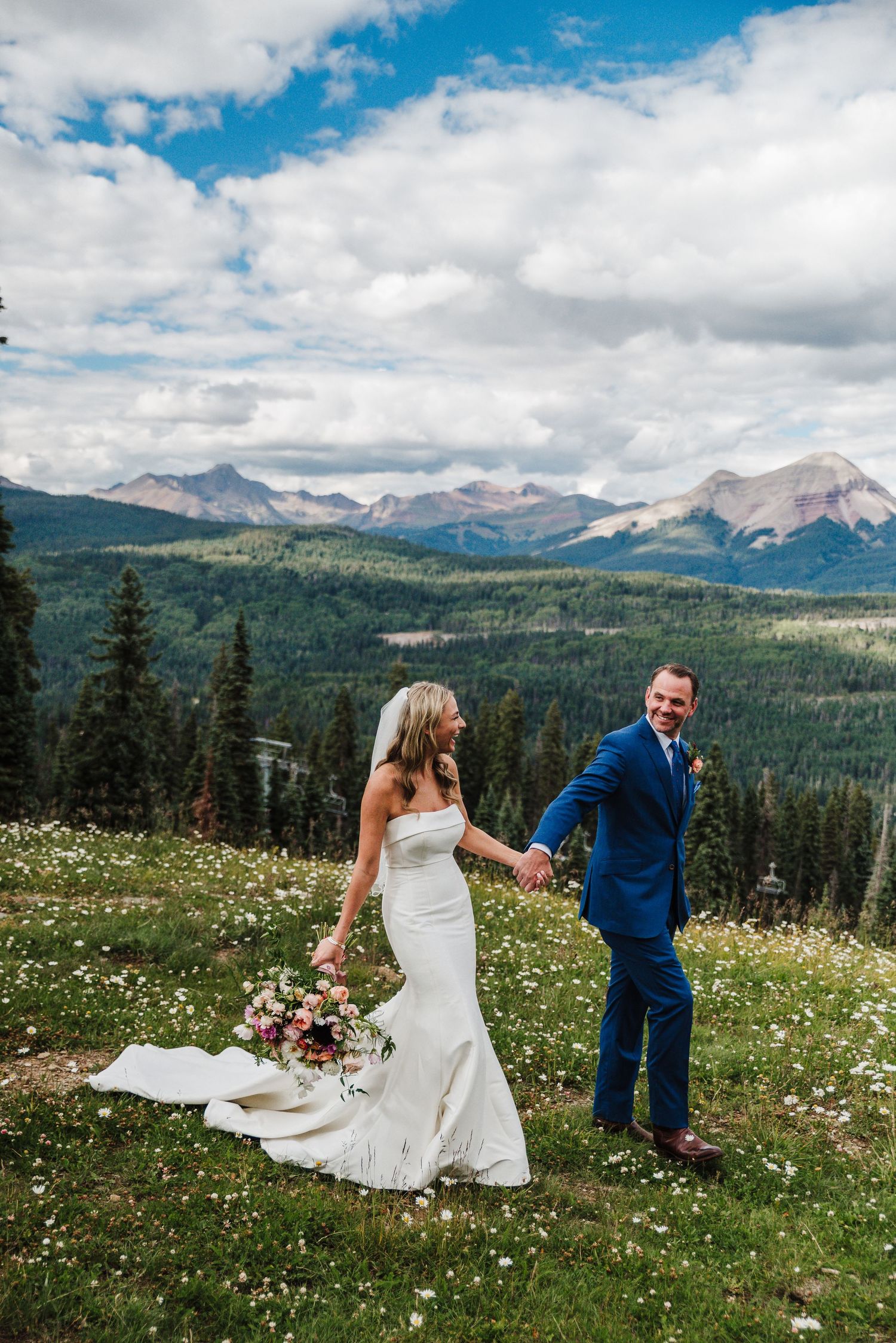 a bride and groom holding hands and walking through a field in front of colorado mountains