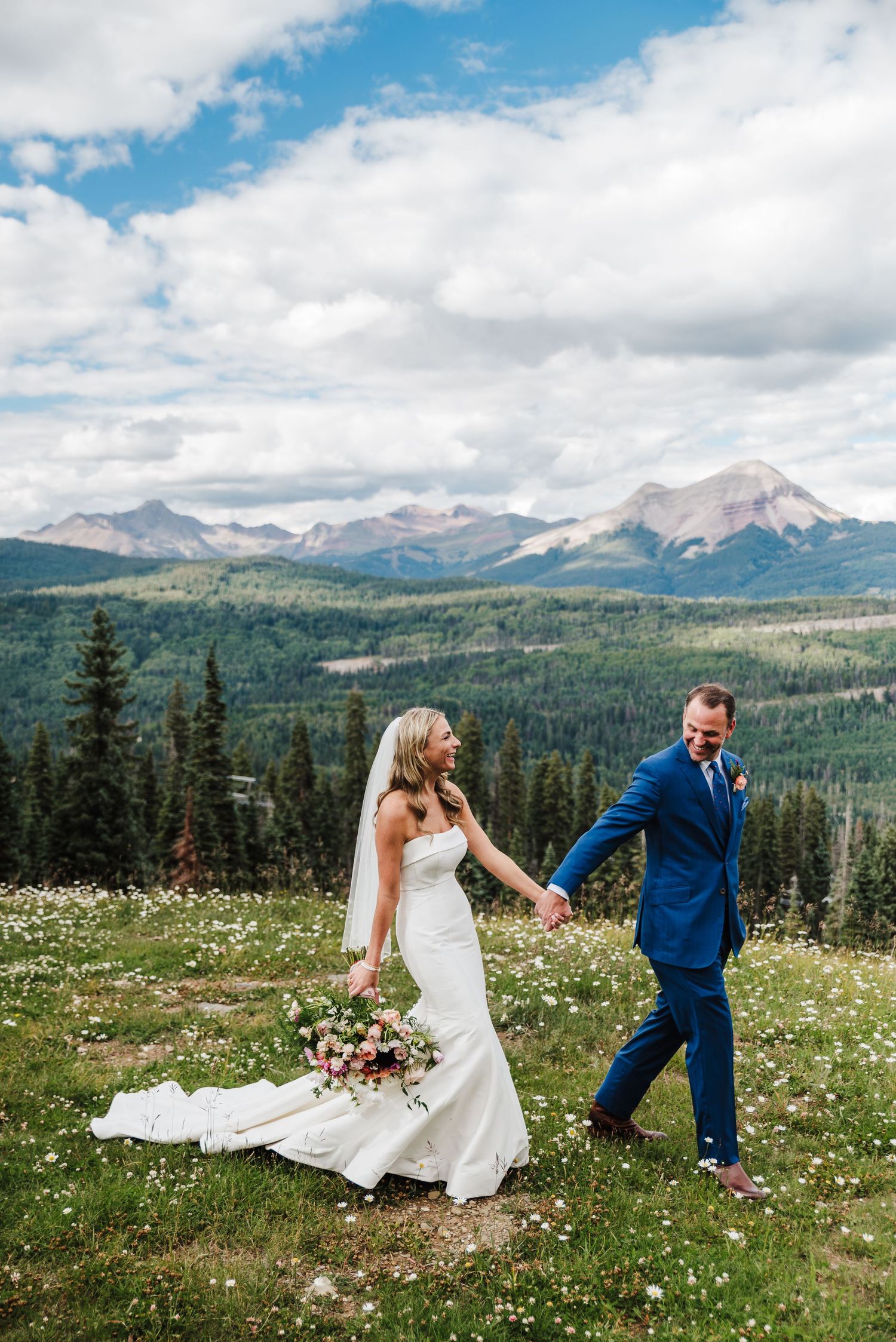 a bride and groom holding hands and walking through a field in front of colorado mountains