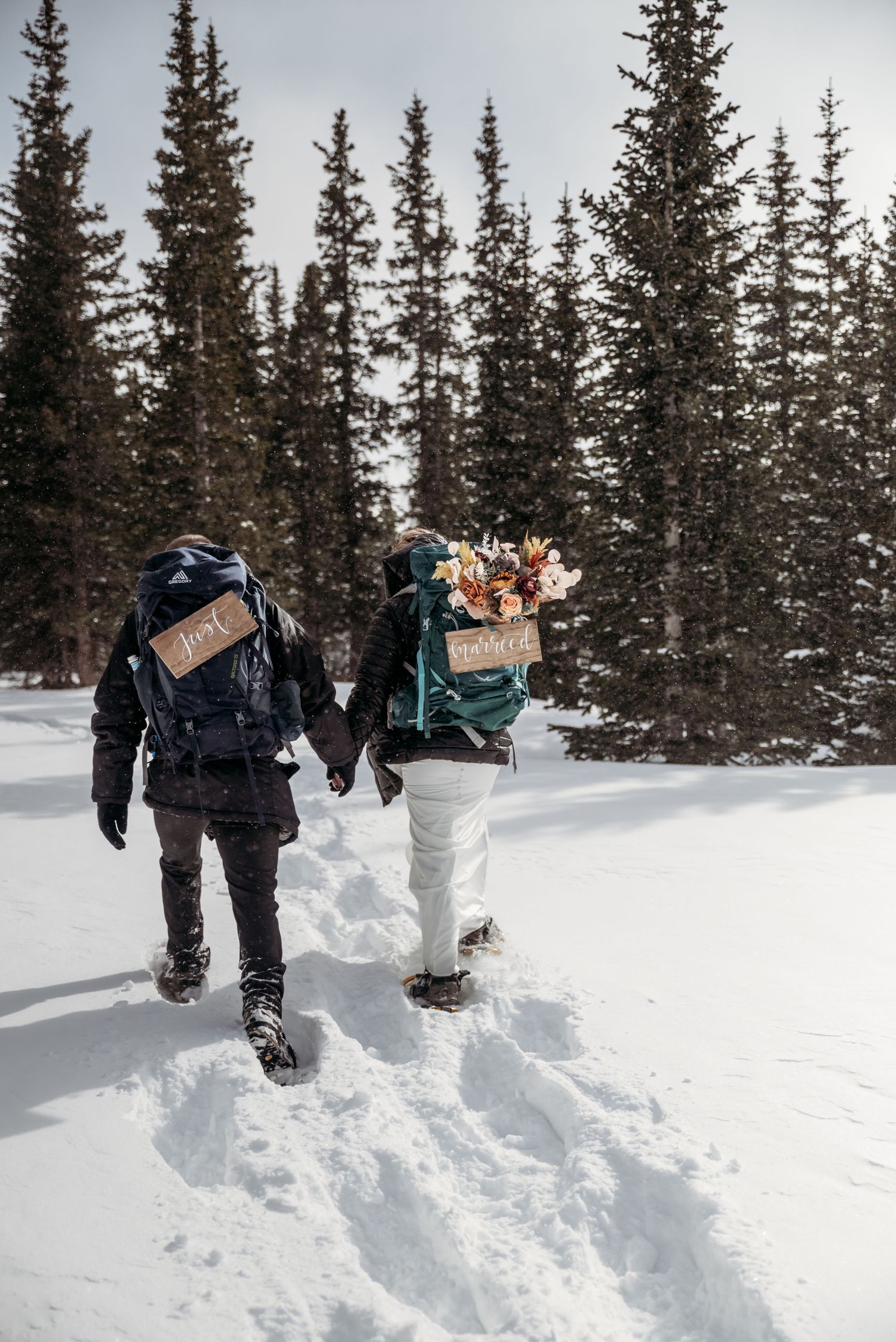 a bride and groom holding hands and snowshoeing through deep snow with backpacks
