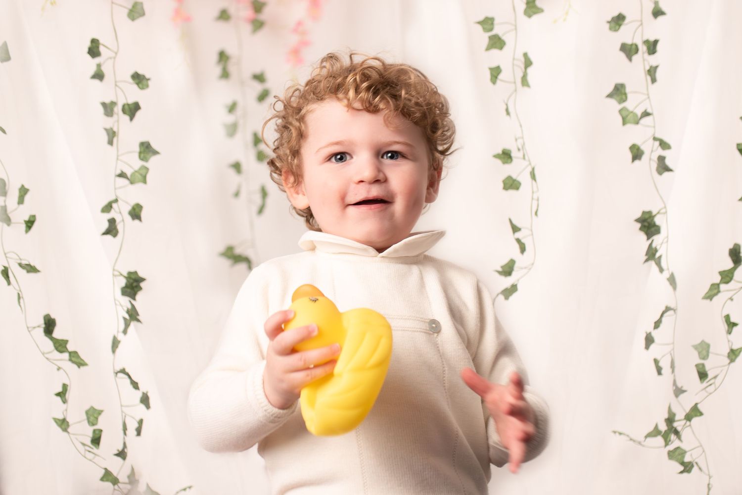 A smiling child in a white sweater holds a plush yellow banana toy against a backdrop with hanging vines.