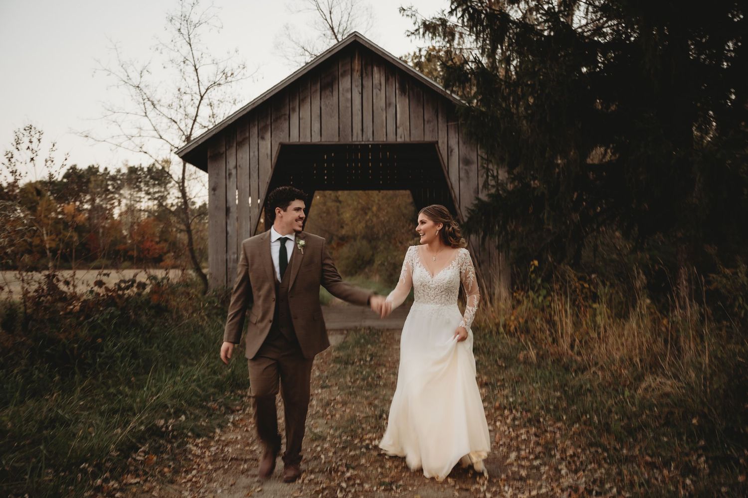 Bride and groom holding hands walking near rustic covered bridge surrounded by autumn foliage.