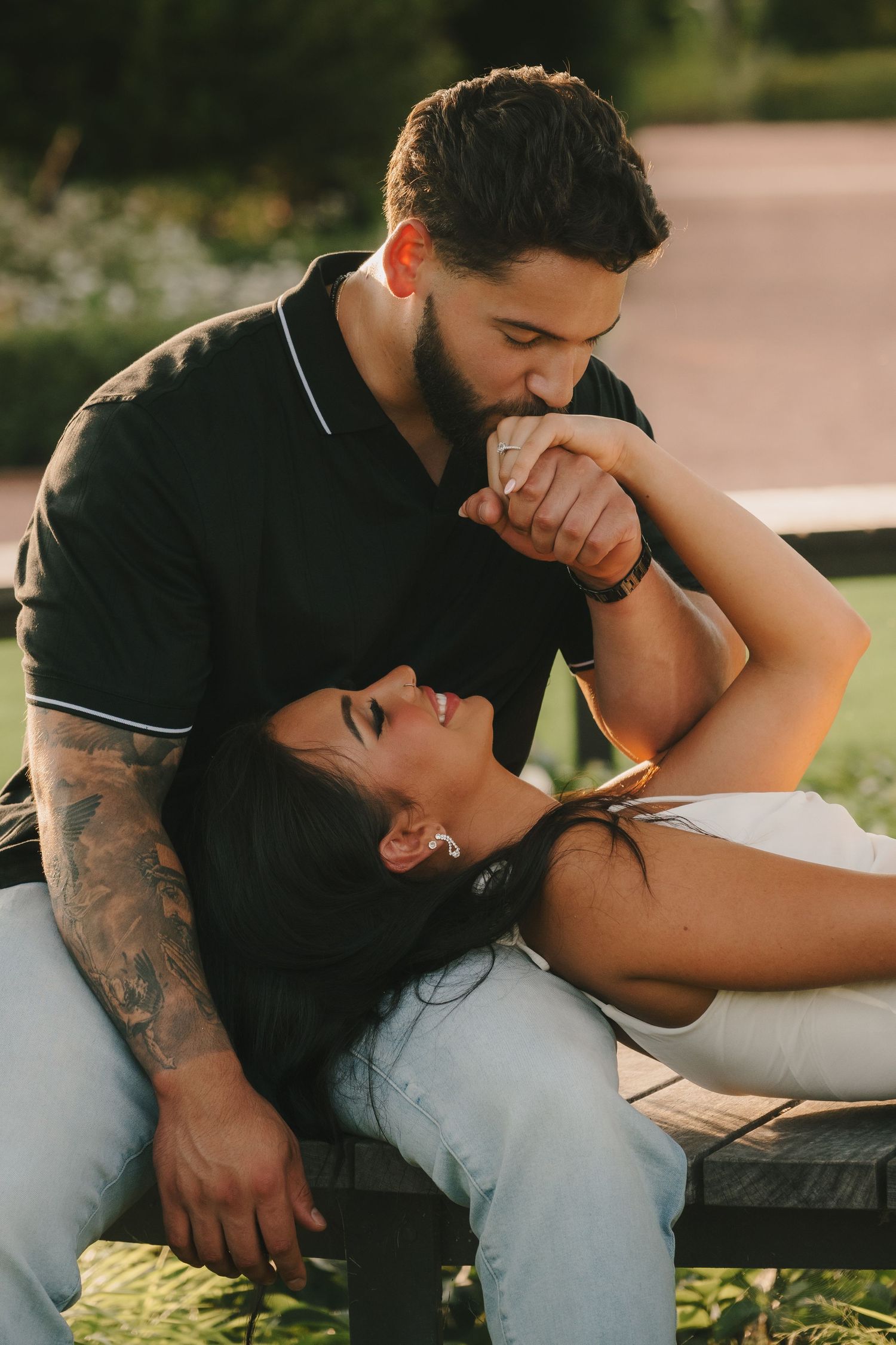 Tattooed man gently kisses his smiling girlfriends hand as she reclines on his lap outdoors at sunset.