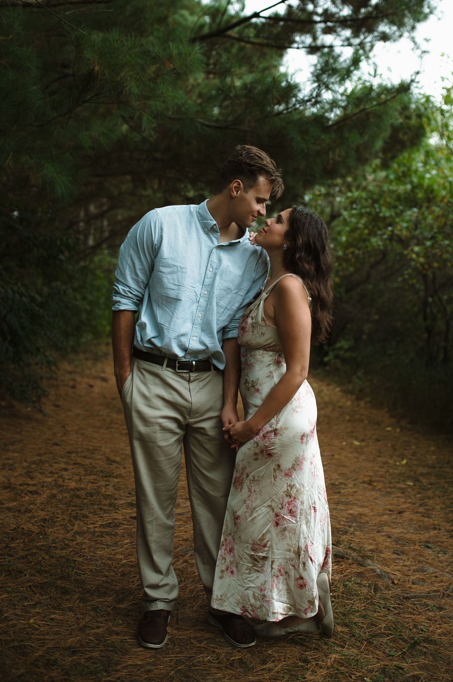 Couple sharing a romantic almost-kiss on a forest path surrounded by tall pine trees.