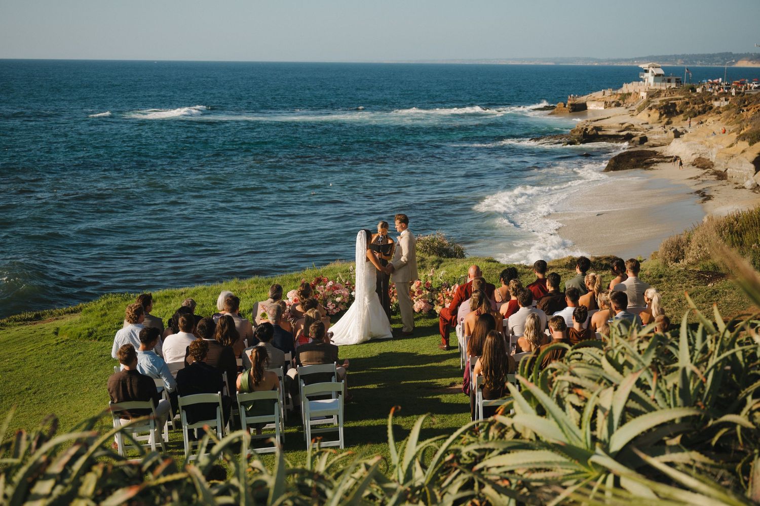 Outdoor wedding ceremony on a cliffside in California overlooking the ocean with guests seated on a lush green lawn.