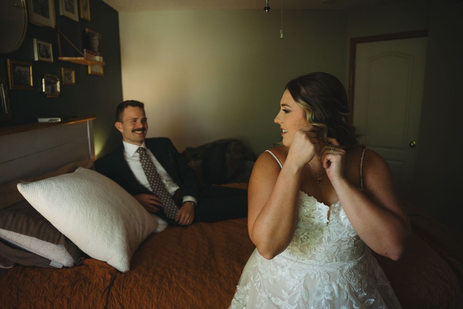 Bride in a lace wedding gown adjusting her earring while groom in suit smiles warmly from a hotel room bed.