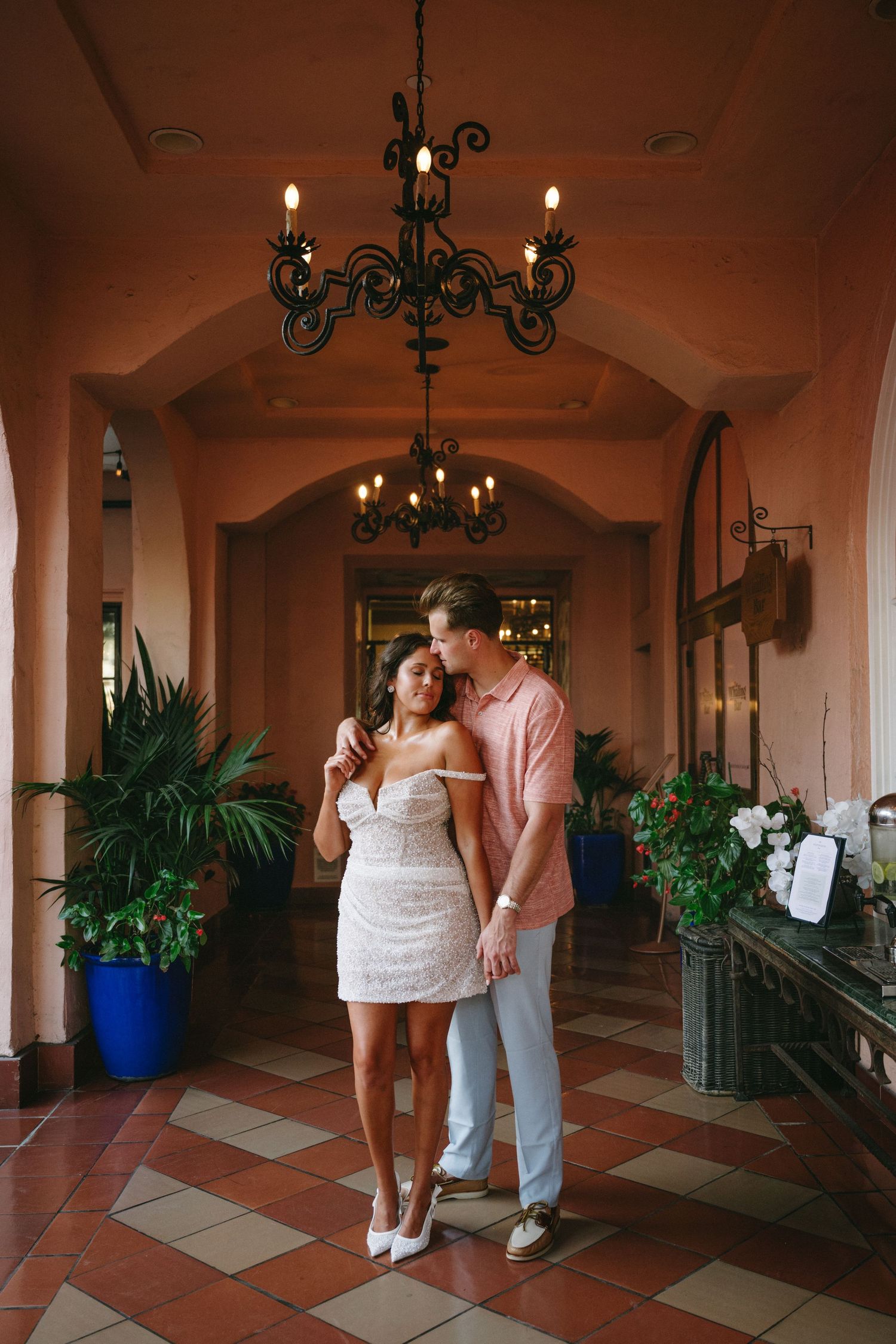Couple embracing in elegant terracotta corridor with chandelier, woman in white dress, man in pink shirt, tropical plants.