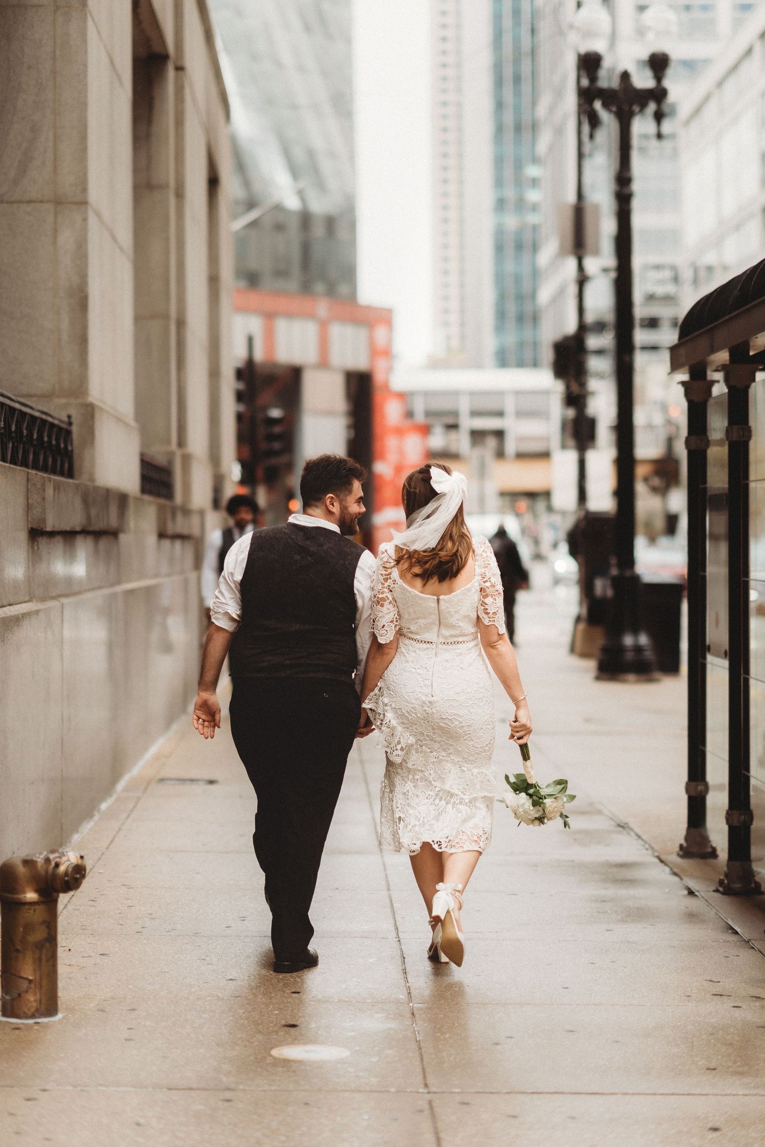 Bride in lace midi dress and groom in vest walking arm in arm down a city sidewalk.