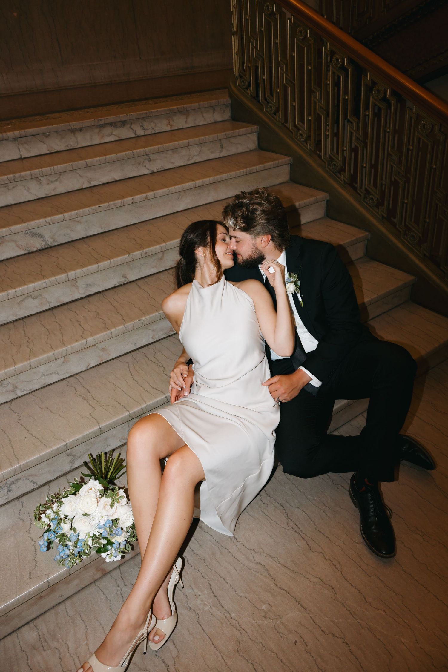 Newlyweds kissing on marble staircase with bouquet of blue and white flowers nearby.