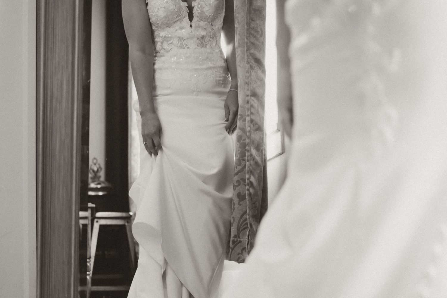 Black and white photo of a bride in a fitted lace wedding gown standing before a mirror.