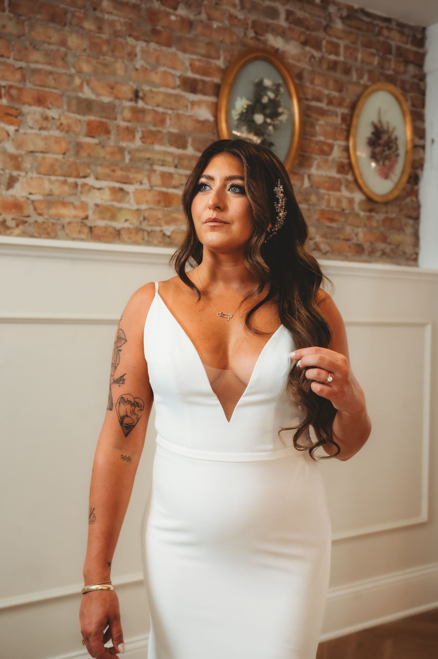Tattooed bride in deep V-neck white gown posing before exposed brick wall with vintage framed artwork.