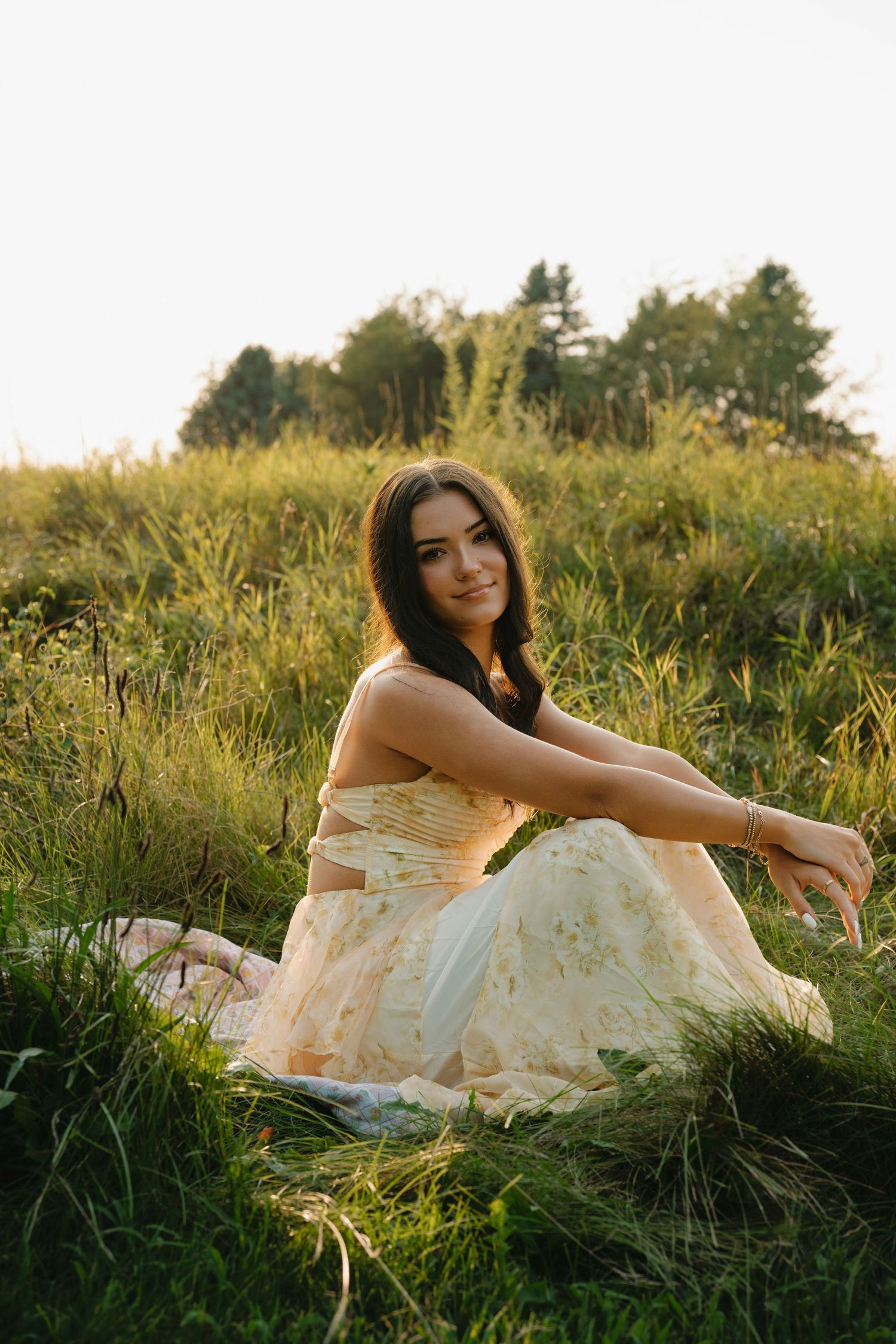 Young woman in floral dress sitting in green meadow, smiling at camera during golden hour sunset.
