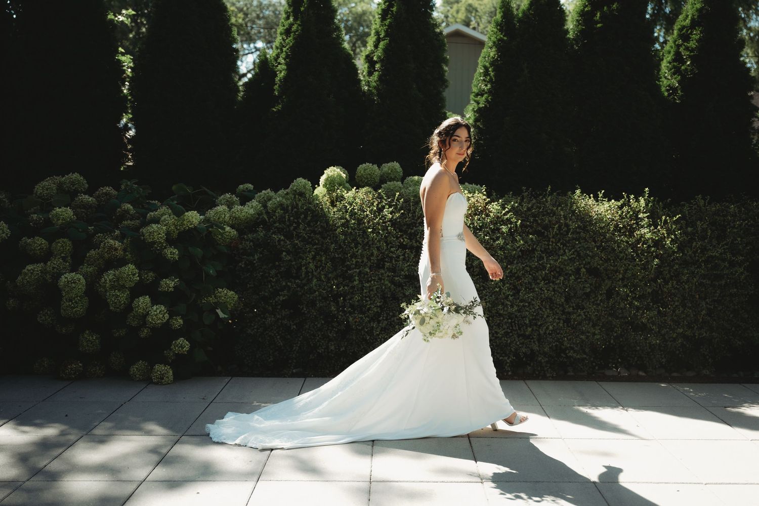 Bride in white gown walking through garden with lush green hedges and hydrangeas holding bridal bouquet.