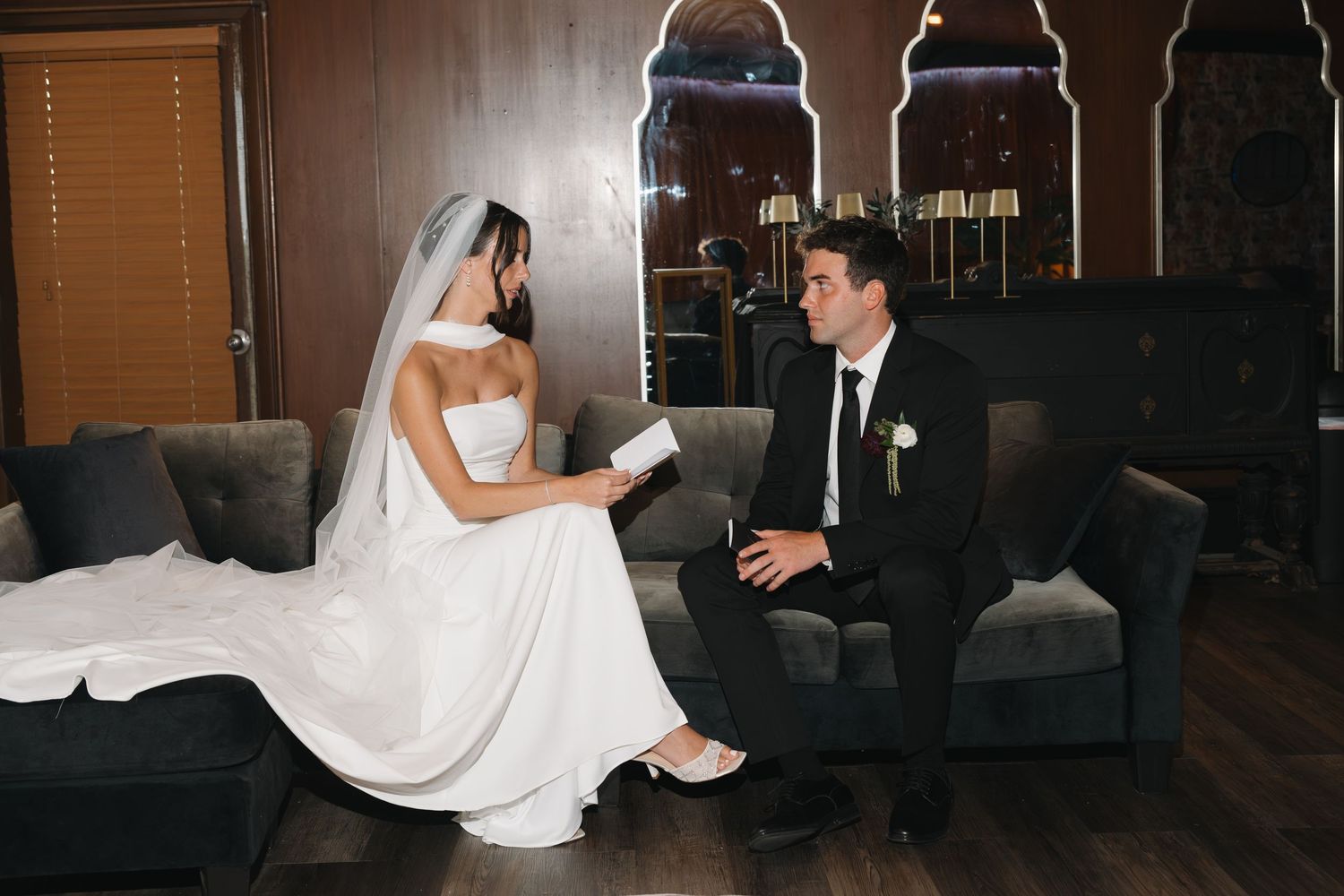 Bride and groom sitting on gray sofa exchanging vows cards in elegant wedding venue.
