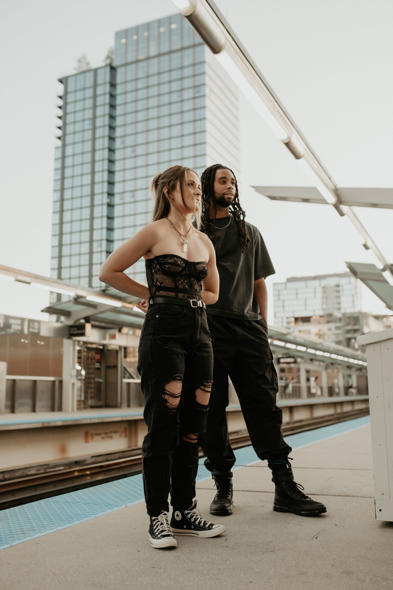Stylish couple posing on urban train platform with city skyscraper in background.