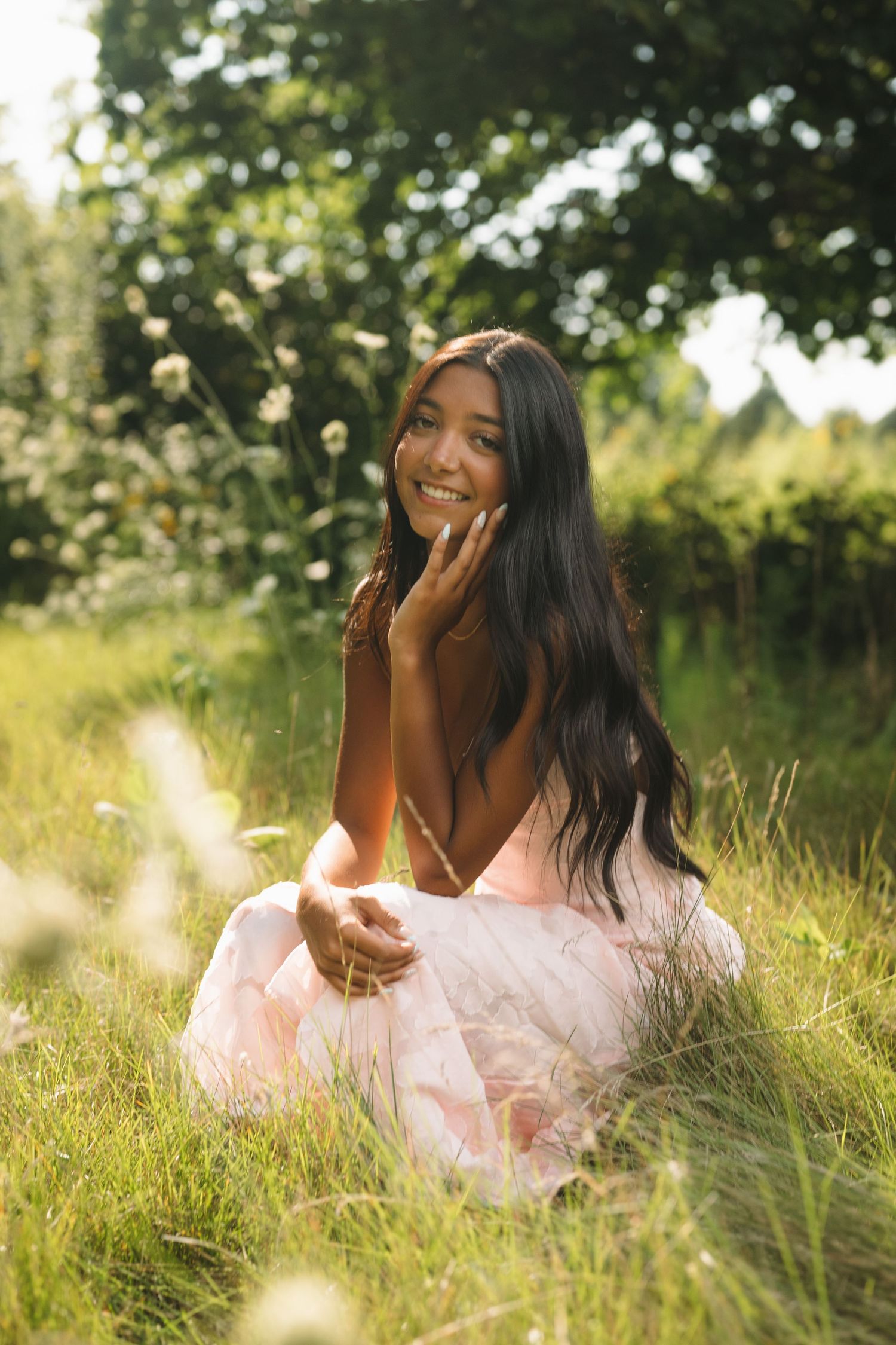 Smiling young woman in pink dress sitting in green grass field with sunlight filtering through trees.