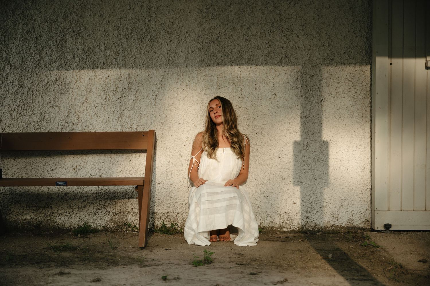 Senior in white dress kneeling gracefully beside a wooden bench against a textured wall in soft light.
