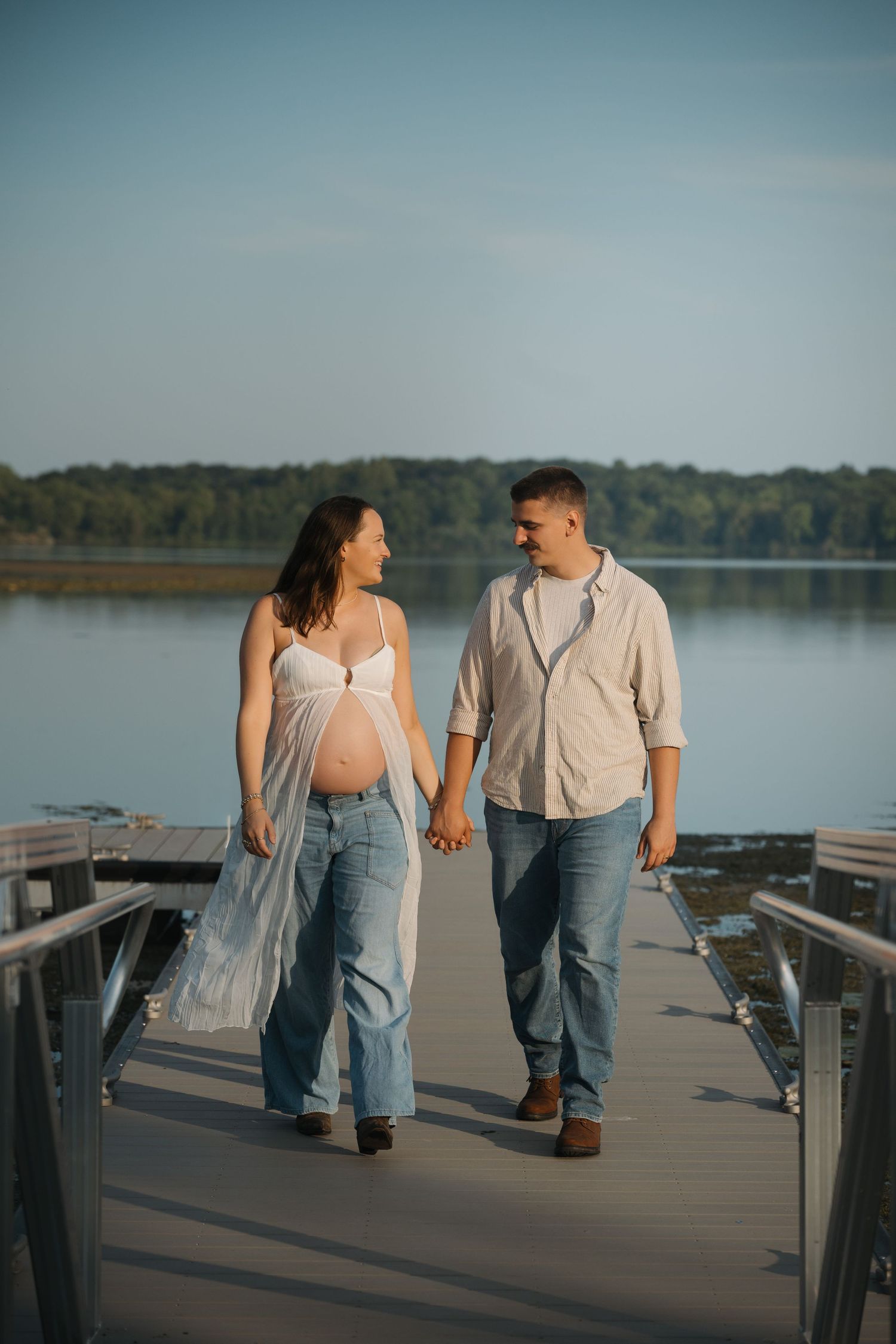 Pregnant couple holding hands on a lakeside dock at sunset, woman in white top and jeans, man in casual shirt.