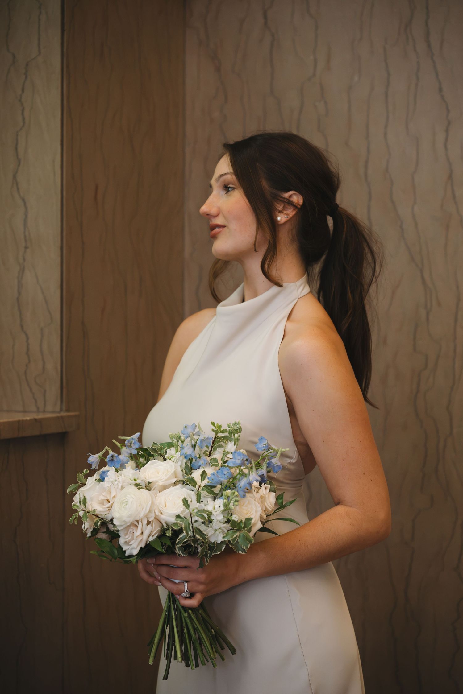 Elegant bride in white halter dress holding blue and white floral bouquet indoors.