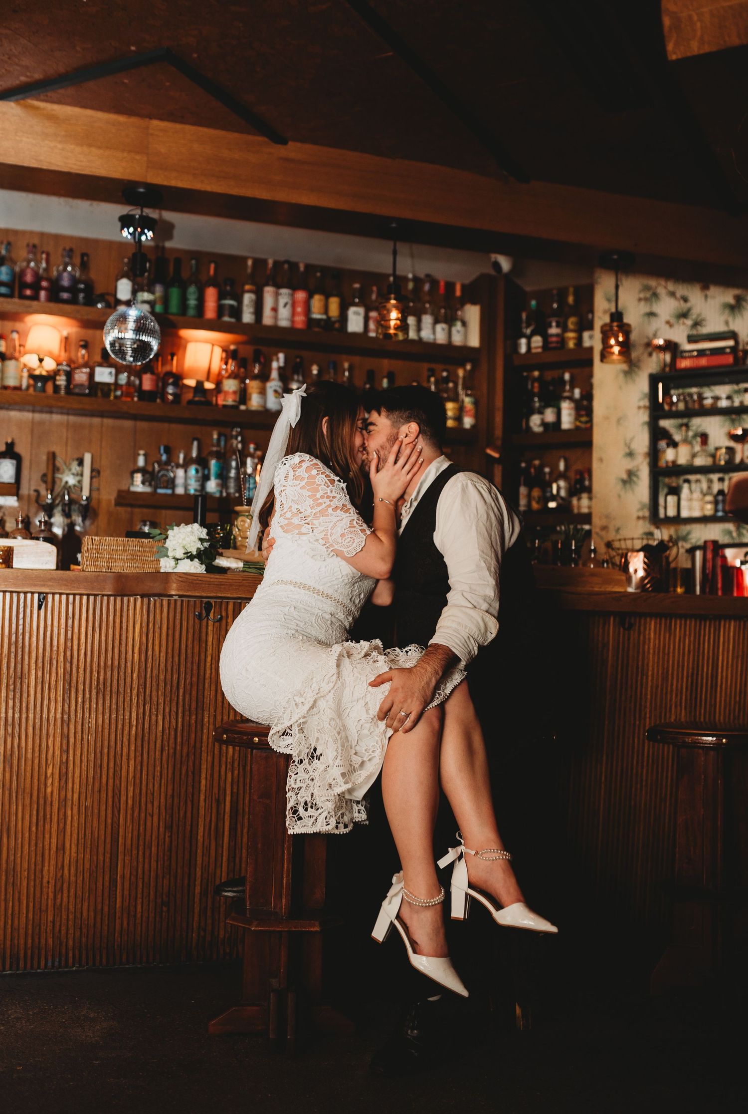 Bride and groom share a passionate kiss at a rustic bar surrounded by bottles of spirits on their wedding day.