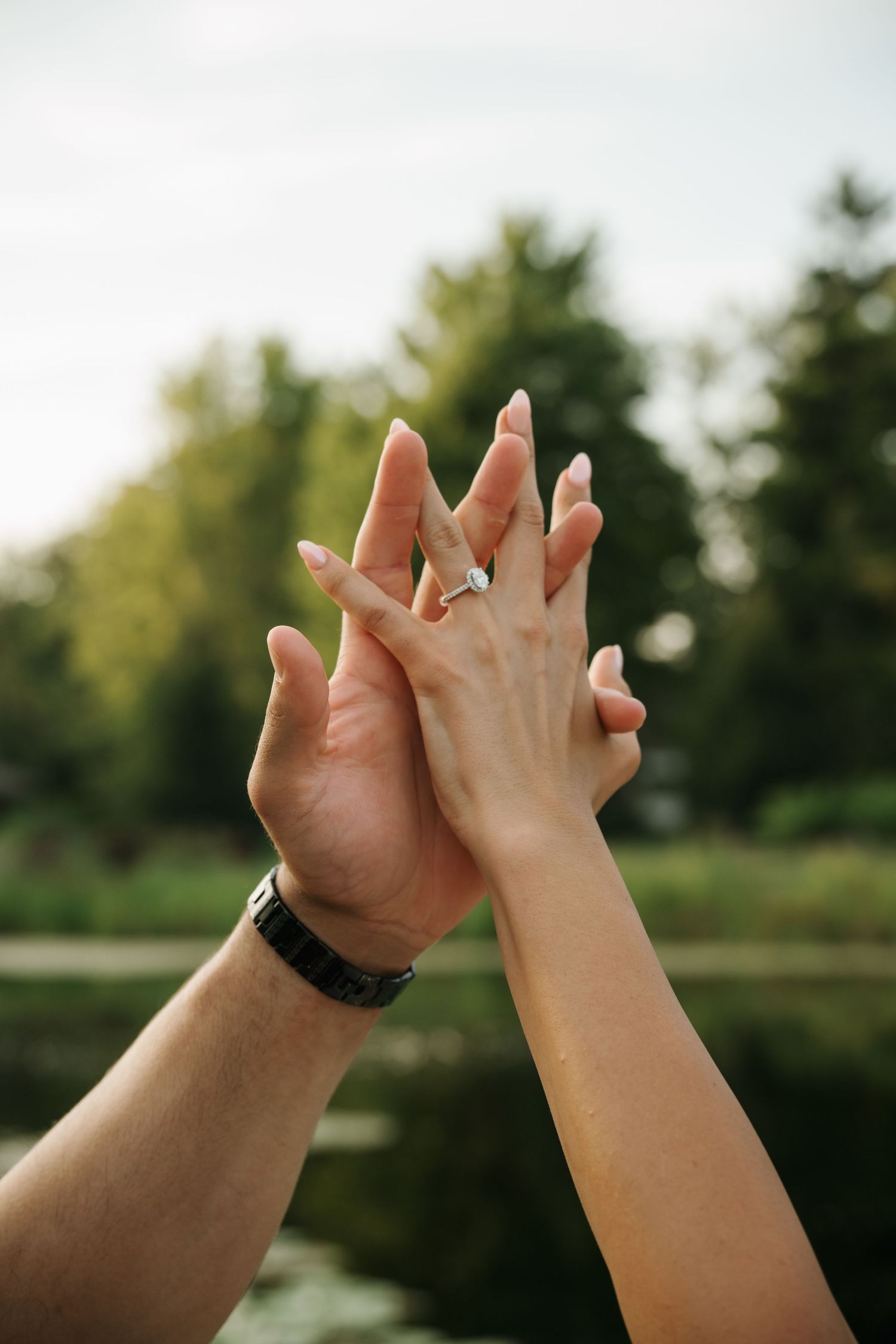 Couple raises intertwined hands showing a sparkling diamond engagement ring against a green outdoor backdrop.